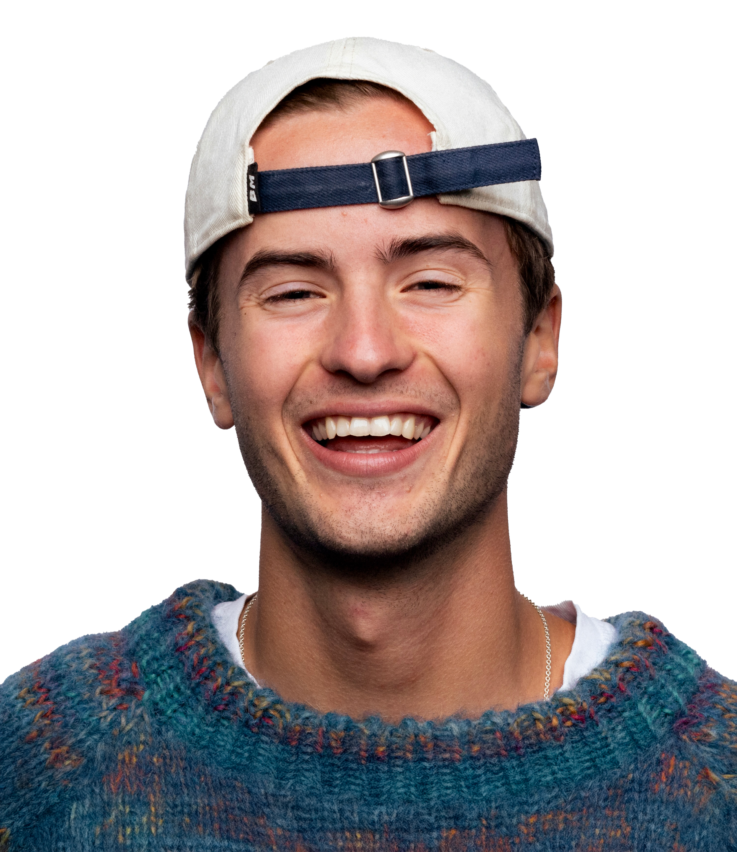 Young man with a baseball cap backwards, smiling, wearing a colorful sweater.