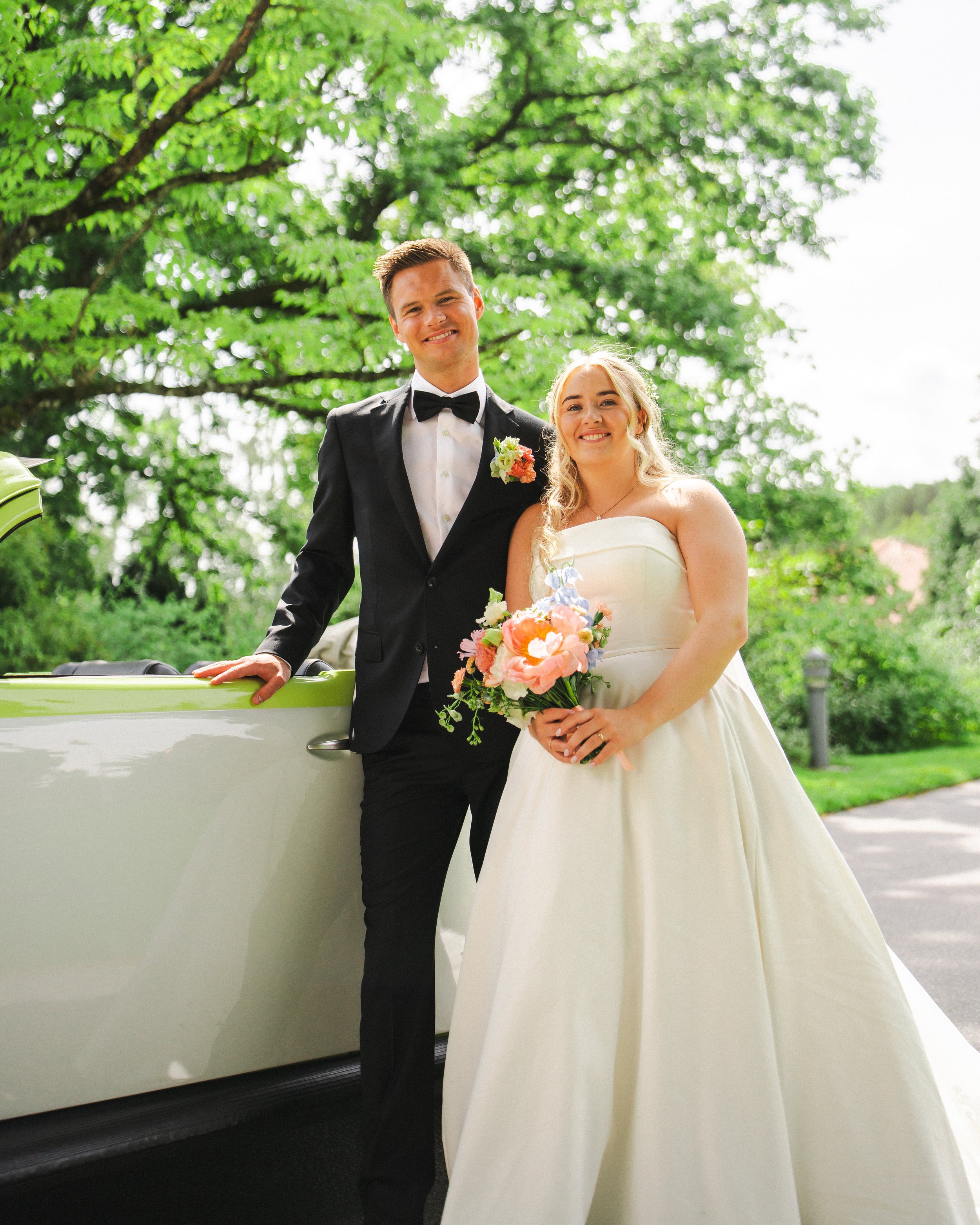 A newlywed couple in wedding attire posing outdoors, with the groom in a black tuxedo and bow tie, and the bride in a strapless white wedding gown holding a bouquet of flowers, standing beside a vintage car with lush green trees in the background.