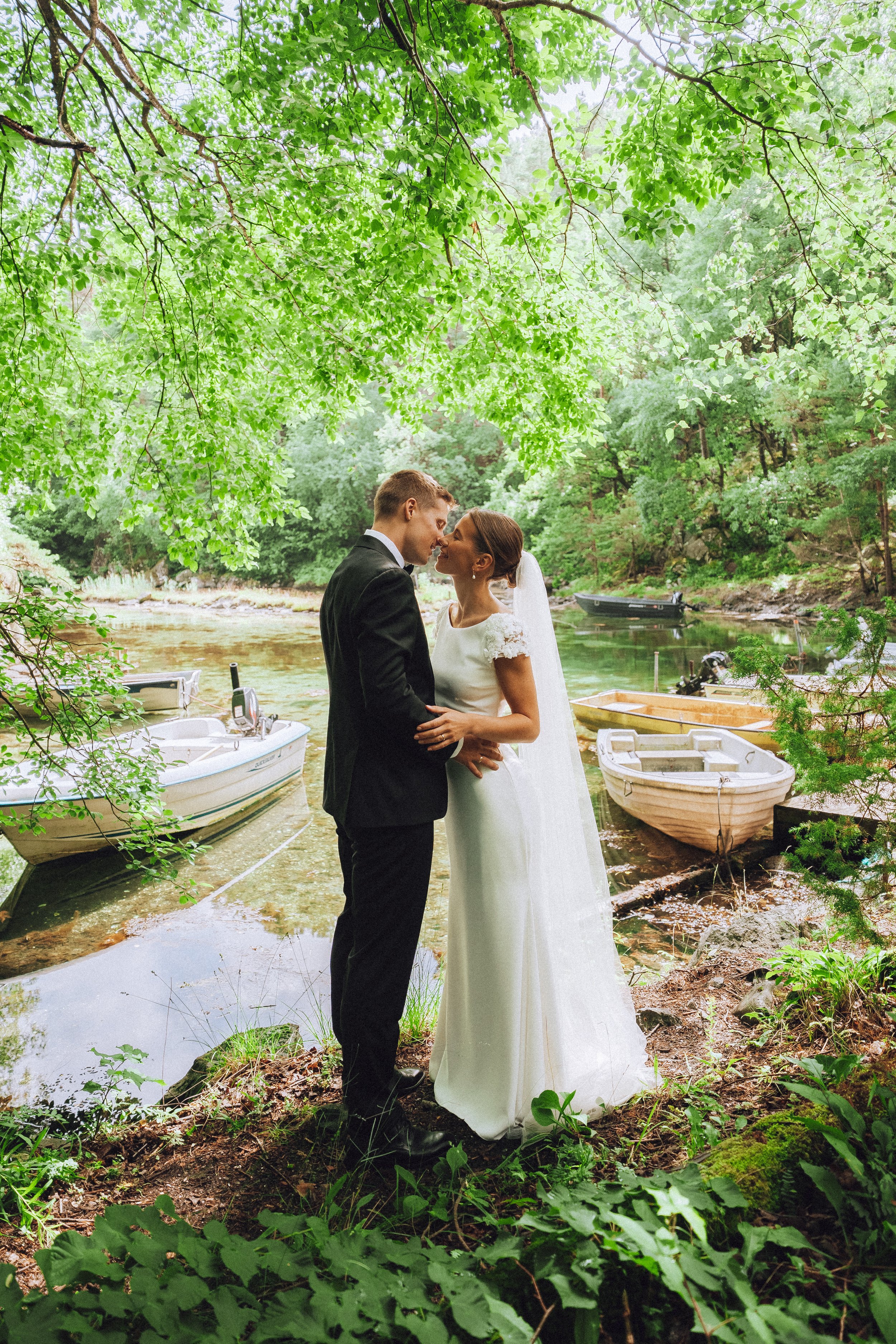 A bride and groom in wedding attire standing close together near a river, surrounded by lush green trees and several boats docked in the water.