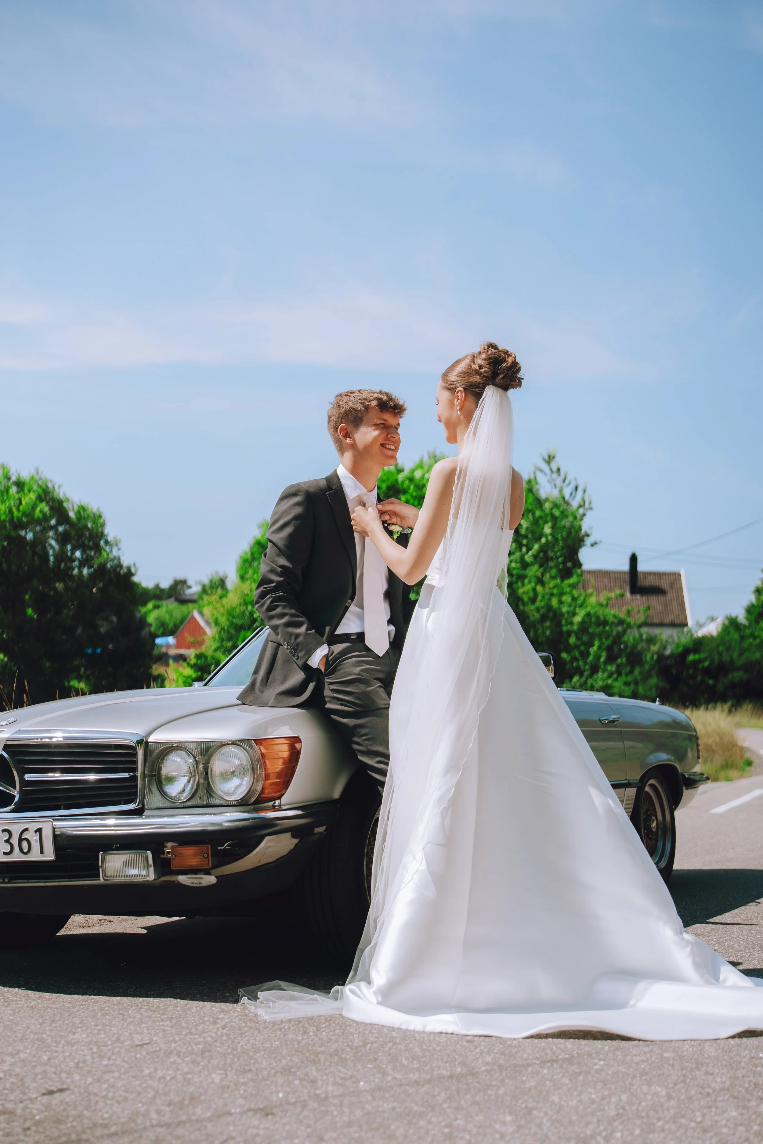 Et brudepar står foran en klassisk Mercedes-bil, hvor bruden i hvit brudekjole og slør holder en hånd på brudgommens skulder, som er kledd i svart dress, på en solrik dag.
