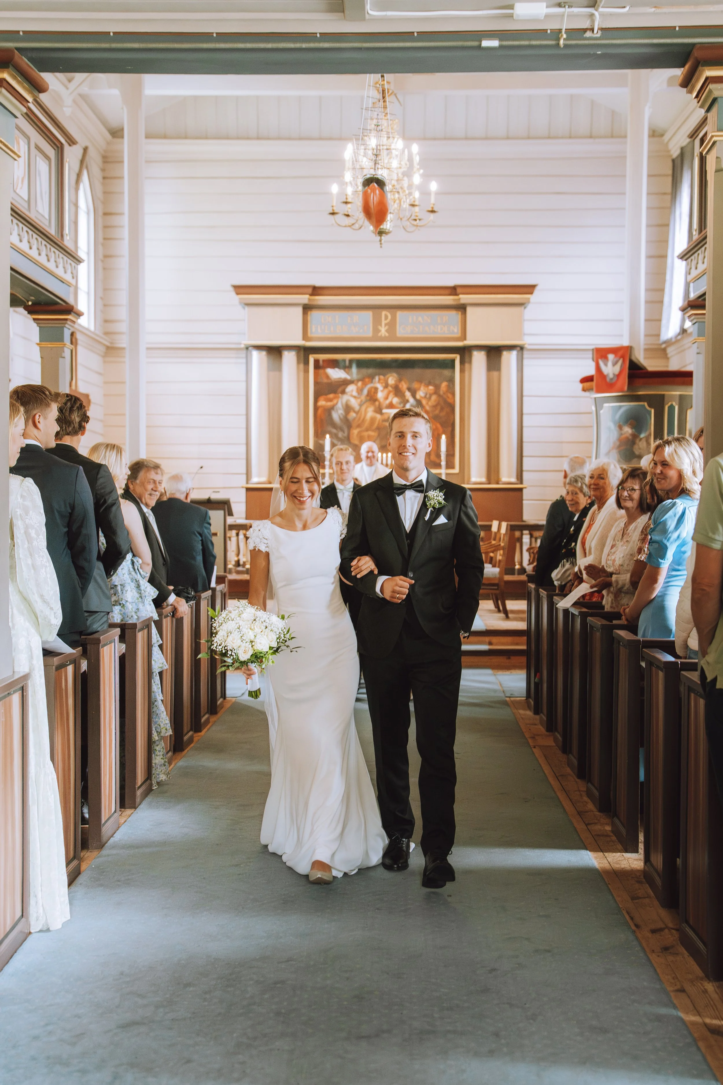 En nygift brud og brudgom går nedover midtgangen i en kirke, omgitt av glade gjester.