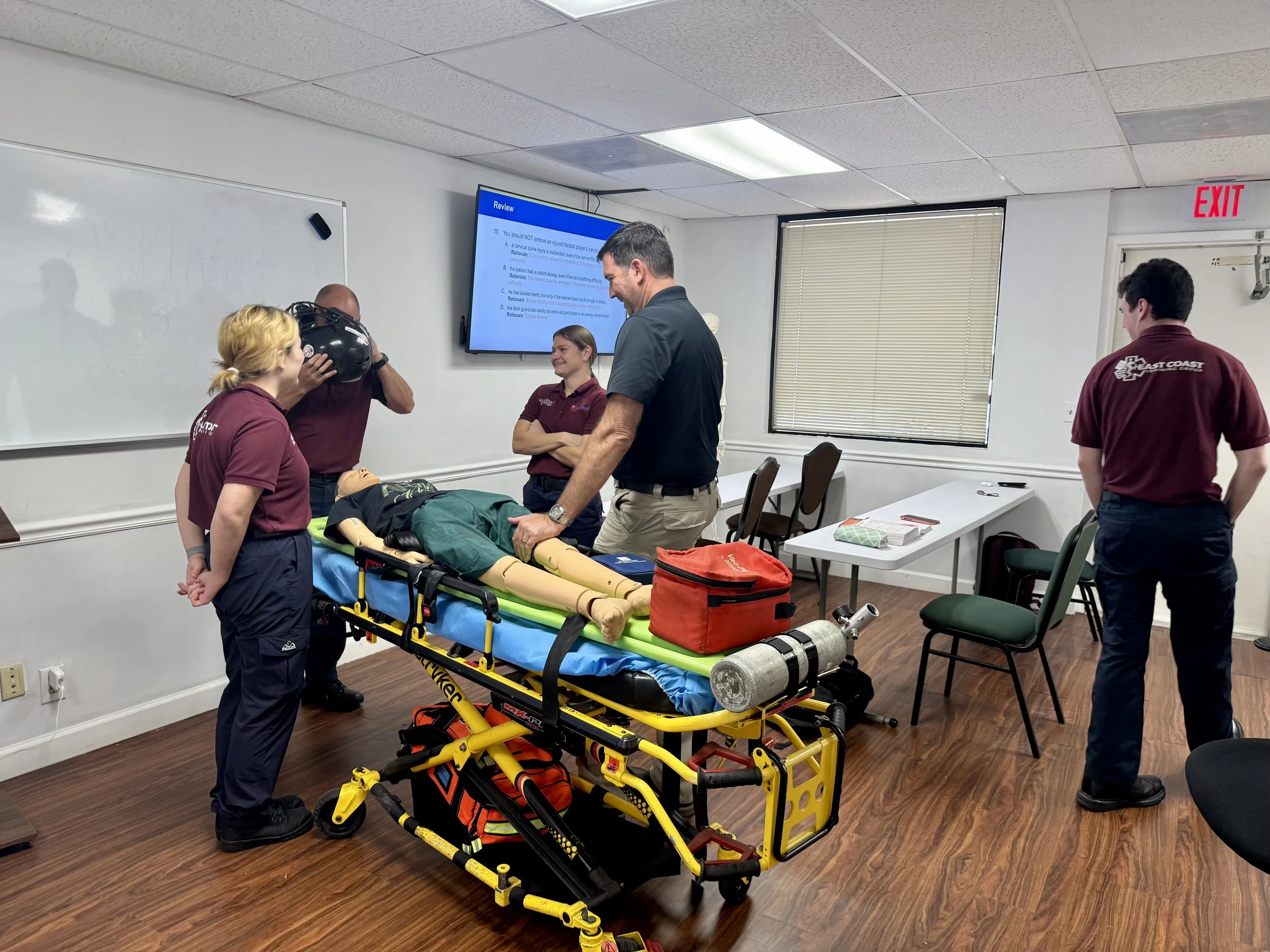A group of people, including emergency responders, are gathered around a medical mannequin on a stretcher in a classroom setting, demonstrating a medical or emergency response training session. There is a large screen with review material on the wall, and various medical equipment and bags are visible.
