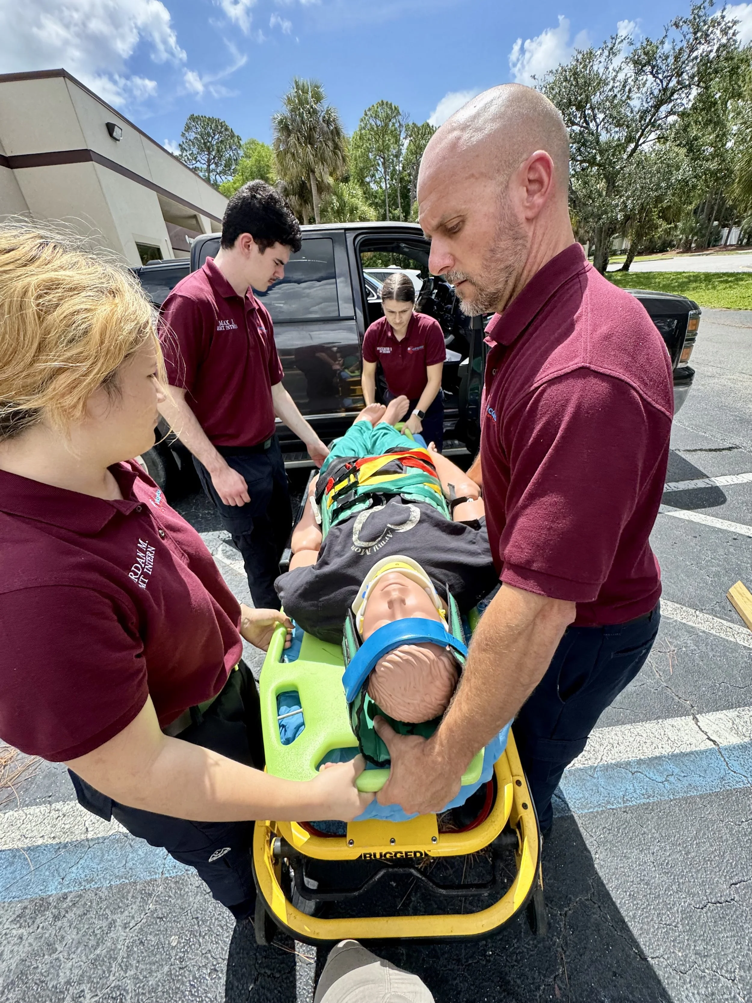 Emergency medical personnel attending to a young patient on a stretcher outside, near an ambulance, under a blue sky with trees in the background.
