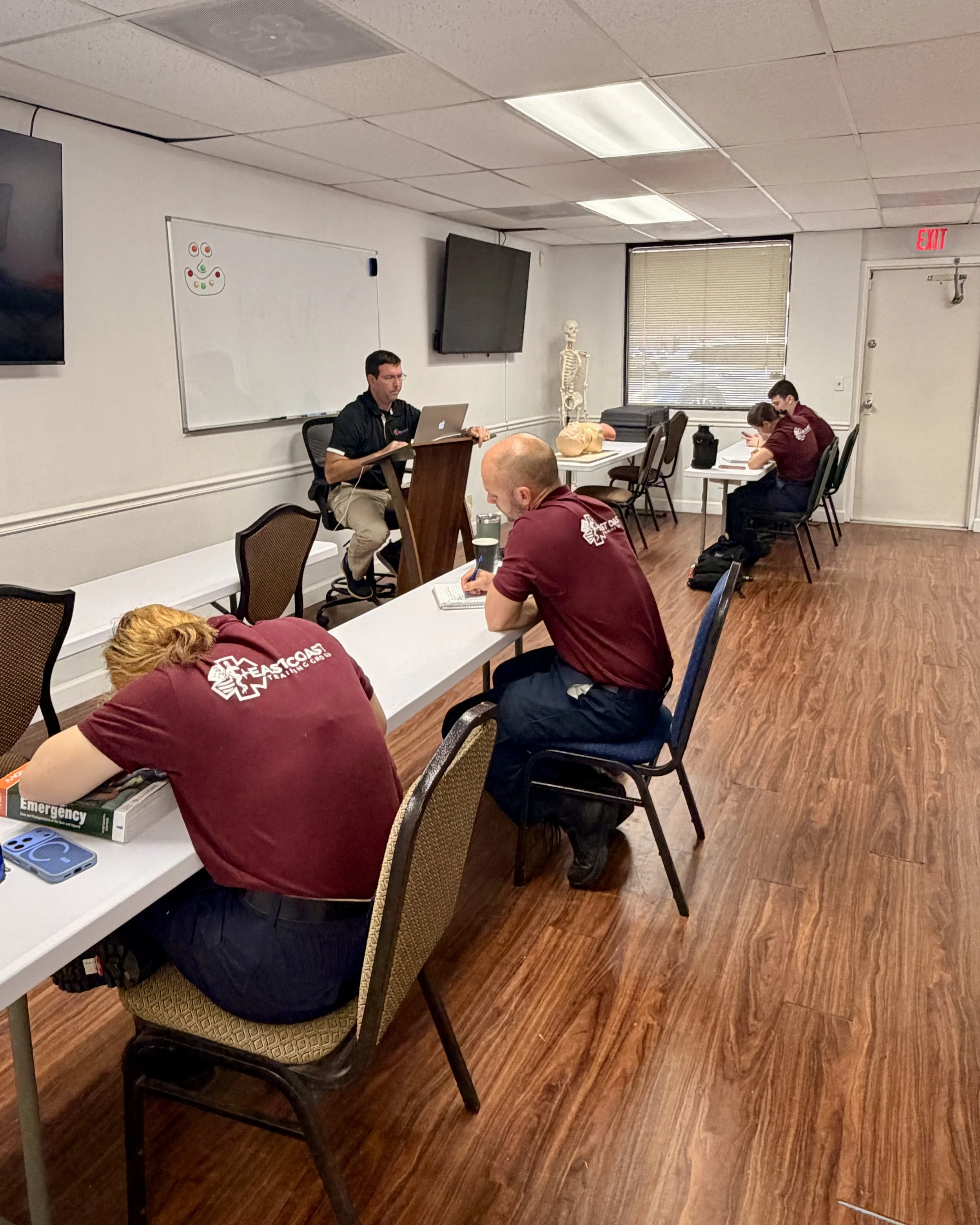 A classroom or meeting room with several people seated at tables. A man is sitting at a small stand with a laptop, and others appear to be taking notes. A skeleton model and a large fake brain are on a table near the window. The room has a whiteboard with some small drawings, a flat screen monitor, and a window with blinds. Some people are wearing matching maroon shirts, and one person has their head down on the table.