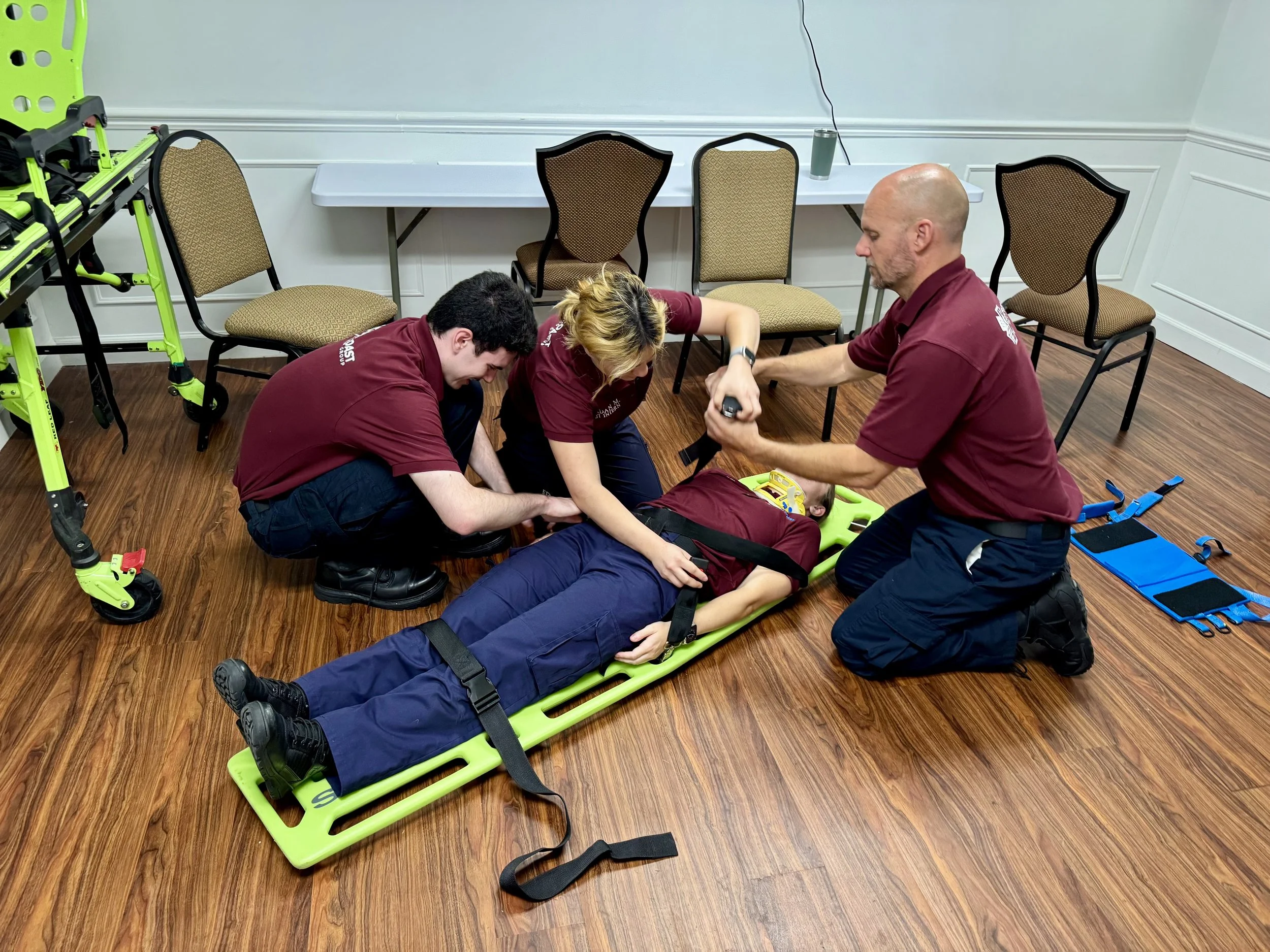 Three emergency responders in matching maroon shirts are attending to a person lying on a stretcher in a room with wooden floors and beige chairs. One responder is kneeling and holding the patient's arm, another is kneeling and examining the patient, and the third is kneeling and assisting. Medical equipment and a walker are visible nearby.