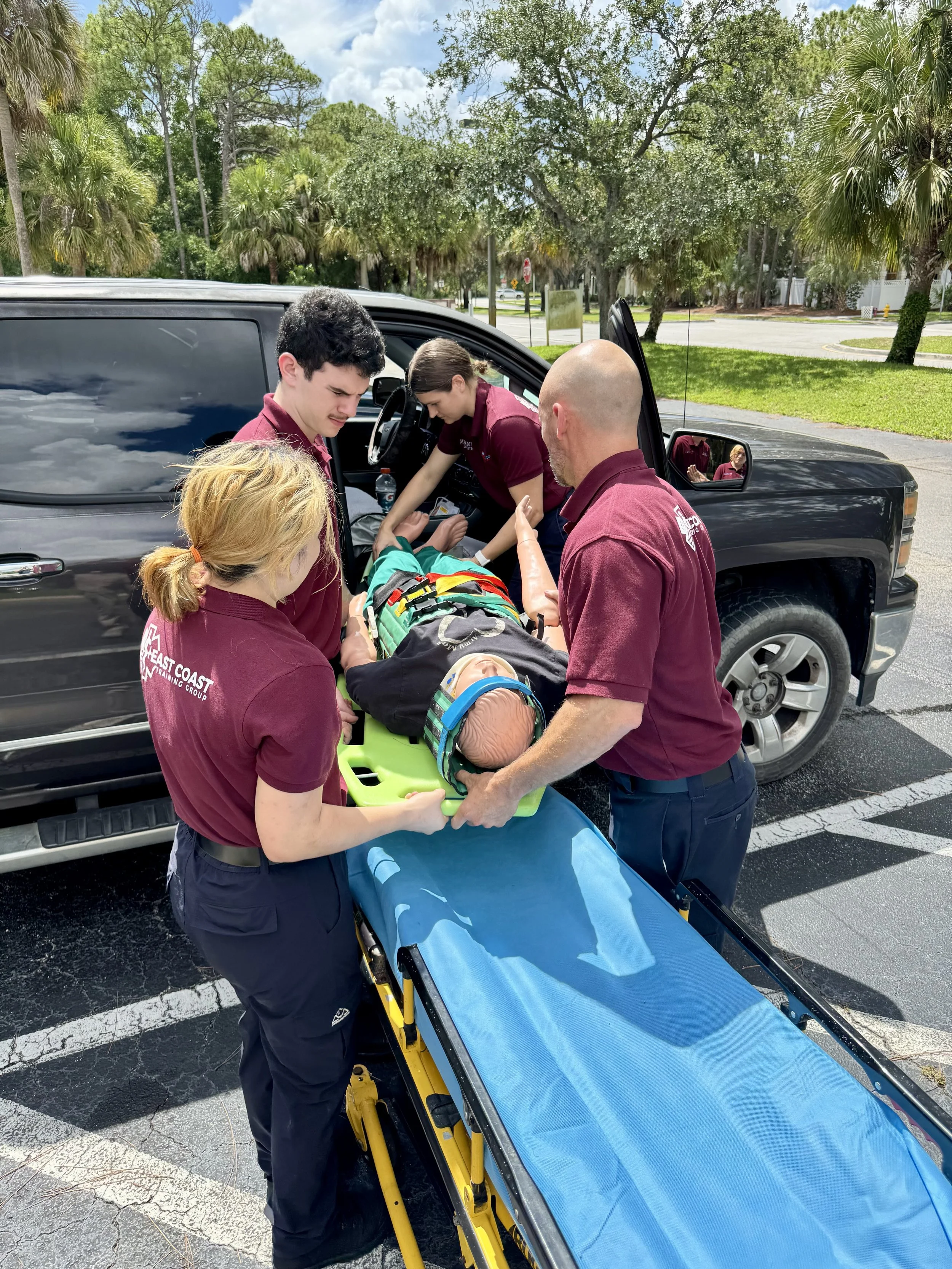 Emergency responders attending to a person lying on a stretcher outside a vehicle in a parking lot on a sunny day with green trees in the background.