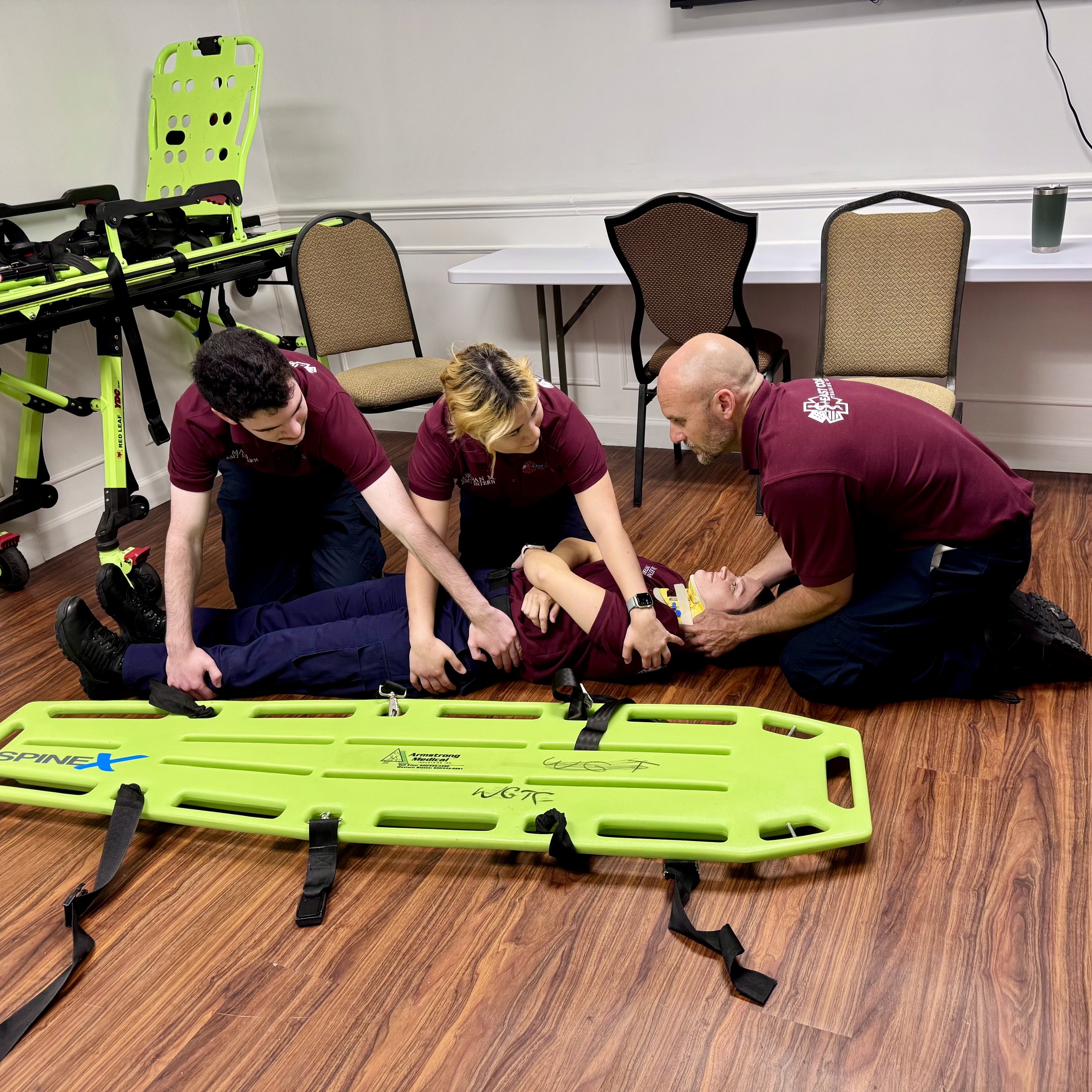 Group of people practicing CPR with an automated external defibrillator (AED) on a person lying on the floor, in a room with chairs, a white wall, and medical equipment.