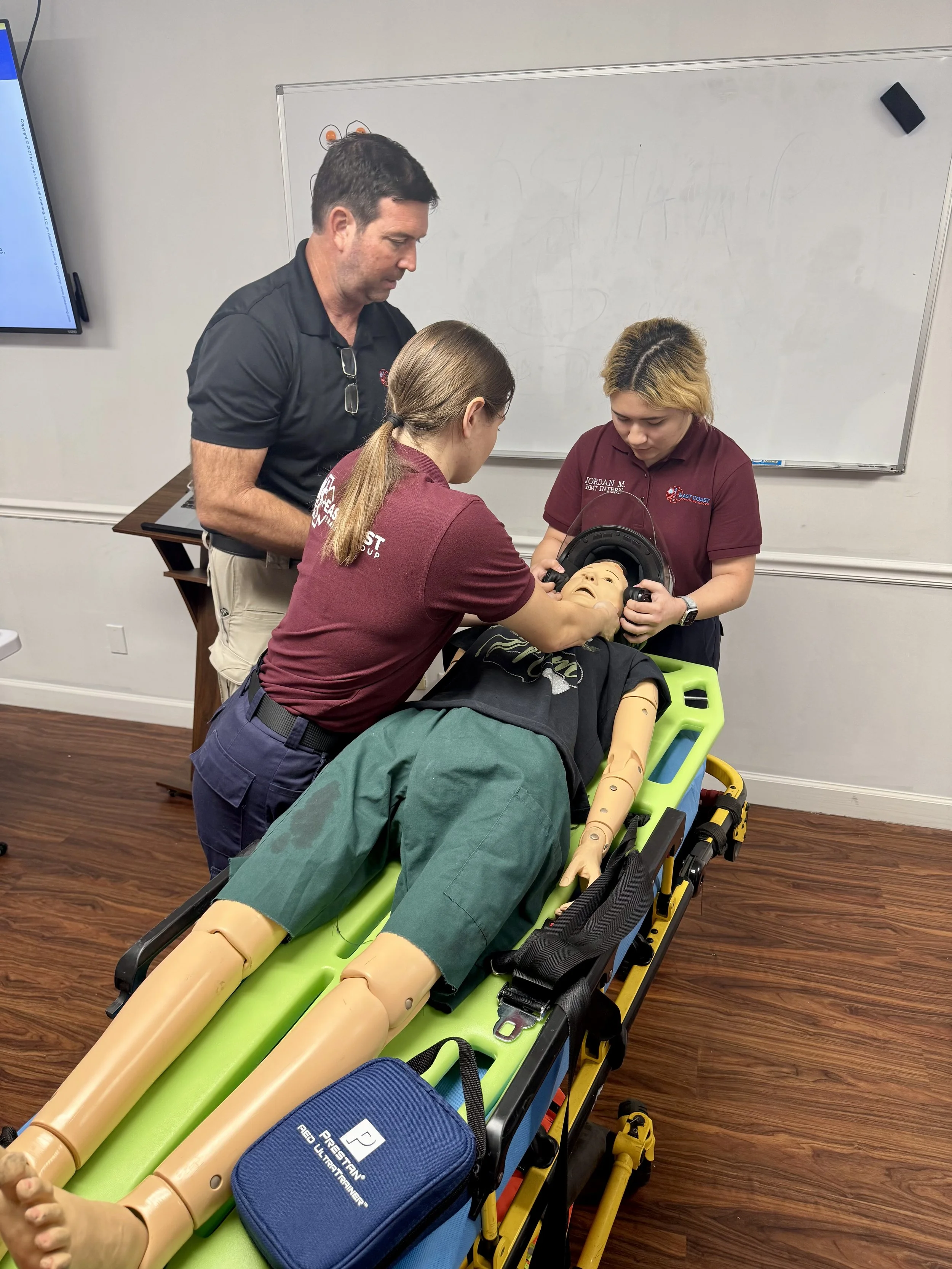Group of emergency responders practicing patient assessment on a medical mannequin lying on a stretcher in a training room with a whiteboard and a monitor.