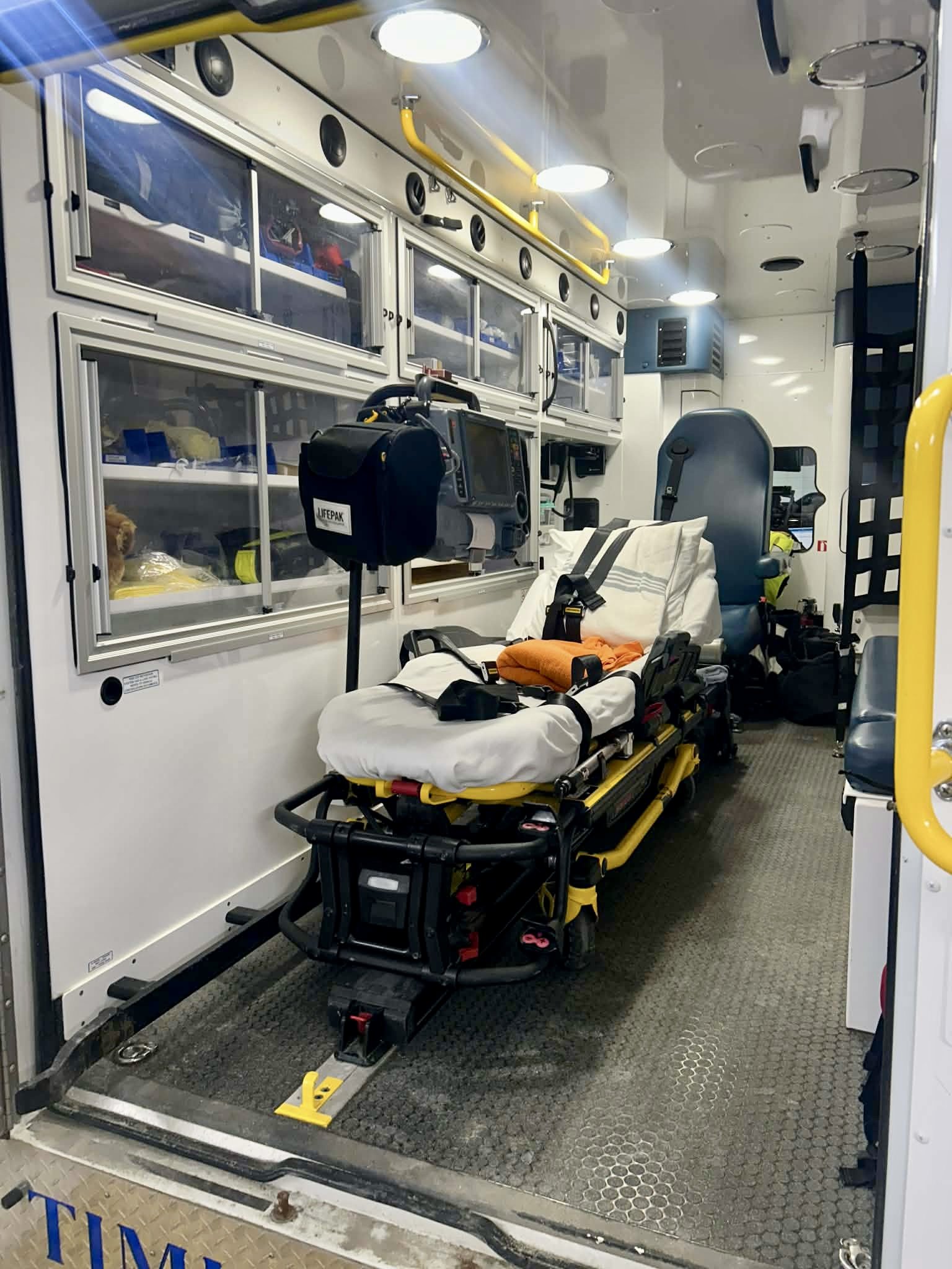 Inside an ambulance with medical equipment, a stretcher with an orange blanket, and various medical supplies and cabinets.