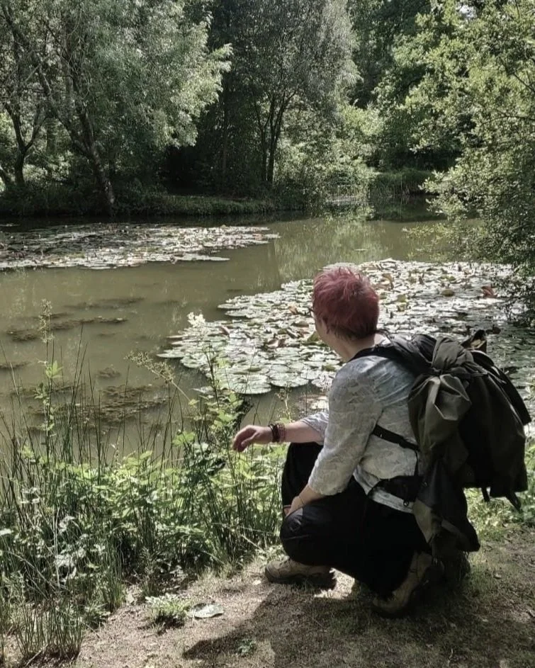Person wearing a backpack crouching by the edge of a pond with lily pads and surrounded by green trees.