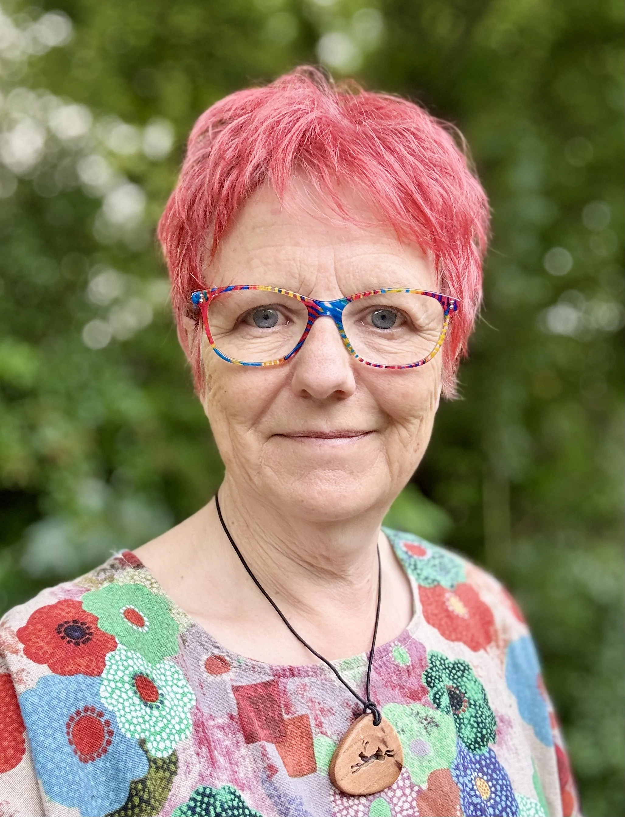 A smiling woman looks straight into the camera. She has short red hair and colorful glasses standing outdoors with greenery in the background. She is wearing a floral patterned top and a necklace with a wooden pendant of a hare.