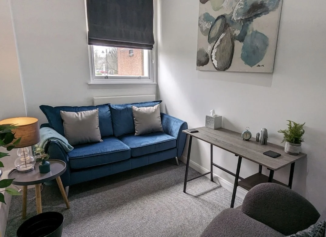 A cozy living room with a blue sofa, gray pillows, a small side table with a lamp, a window with a dark roller shade, a wooden console table with decor, a potted plant, and abstract wall art.