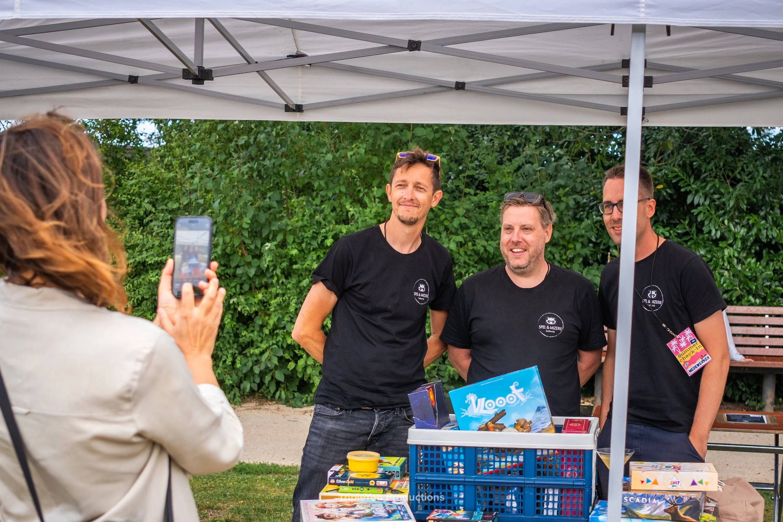 Drie mannen achter een tafel met gezelschapsspelletjes, onder een witte tent, een vrouw die een foto neemt van de mannen voor een groene achtergrond met bosjes en planten