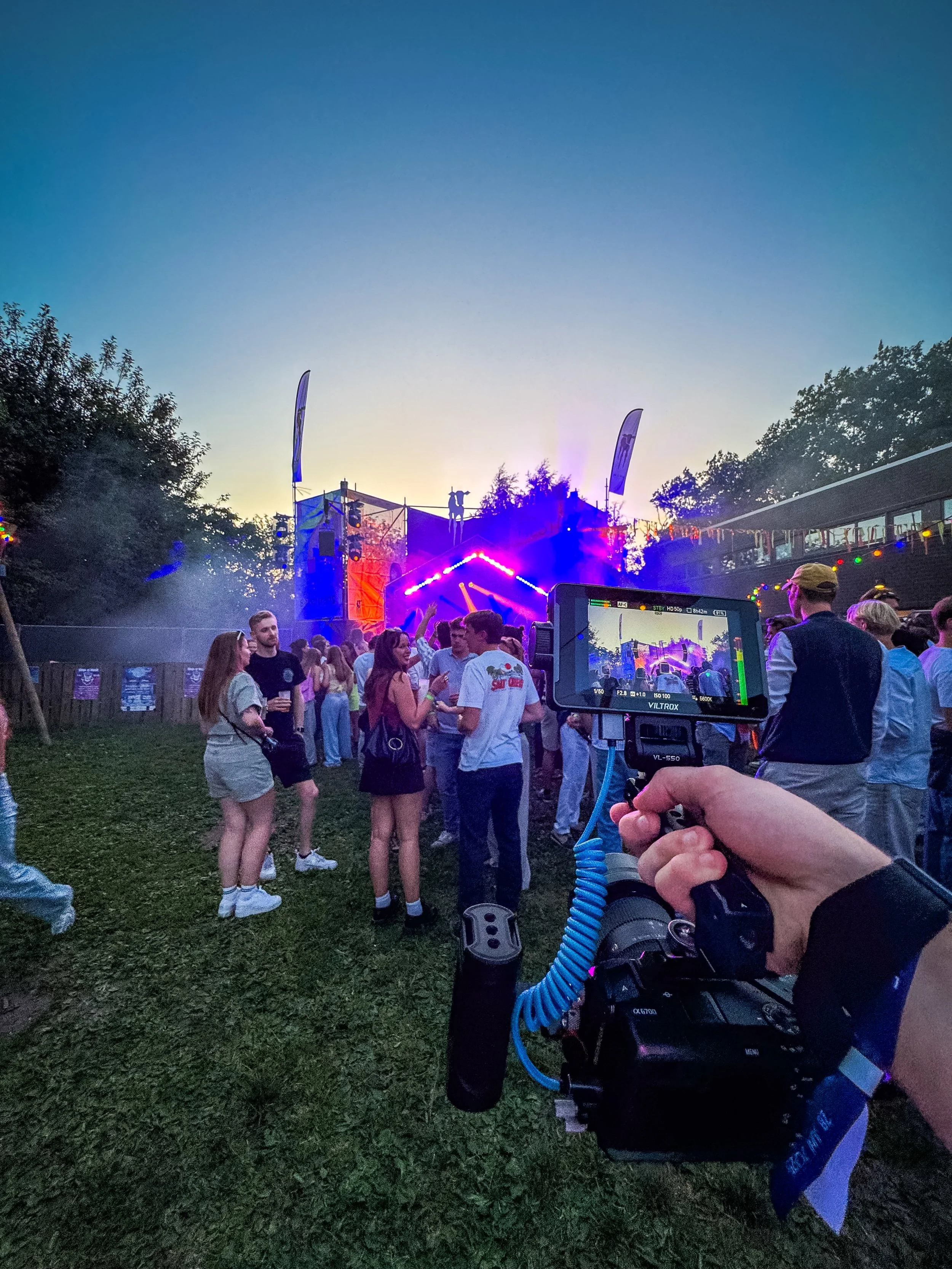Een cameraman die een evenement filmt bij zonsondergang, met een jong publiek voor hem, die aan het dansen en praten zijn, voor een goed belicht podium met moving heads. Errond is een groene omgeving met bomen.