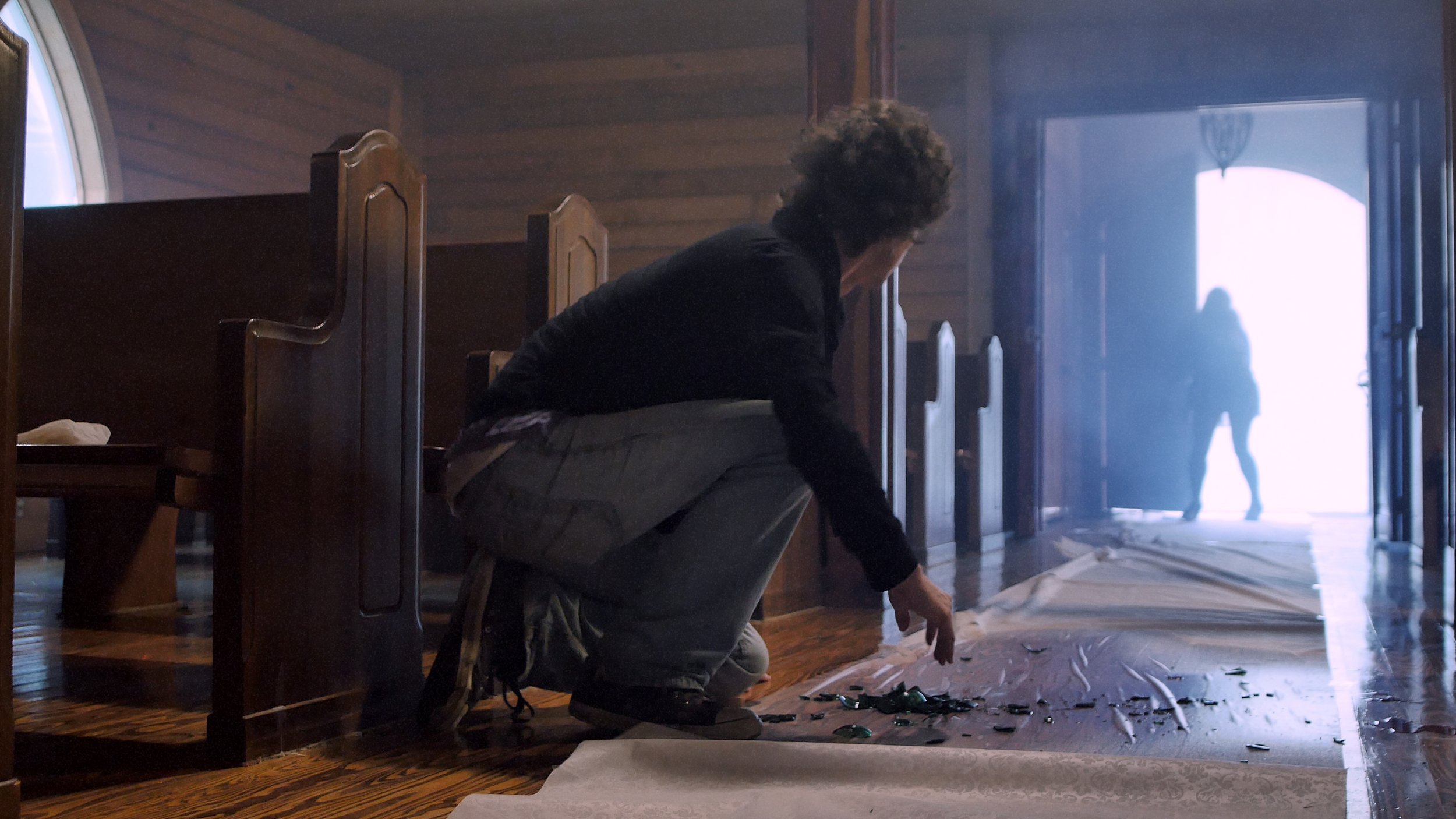 DEAD SATURDAY screen capture: A person crouching inside a church near broken glass on the floor, looking towards the open door where a silhouette of another person is visible outside in bright light.
