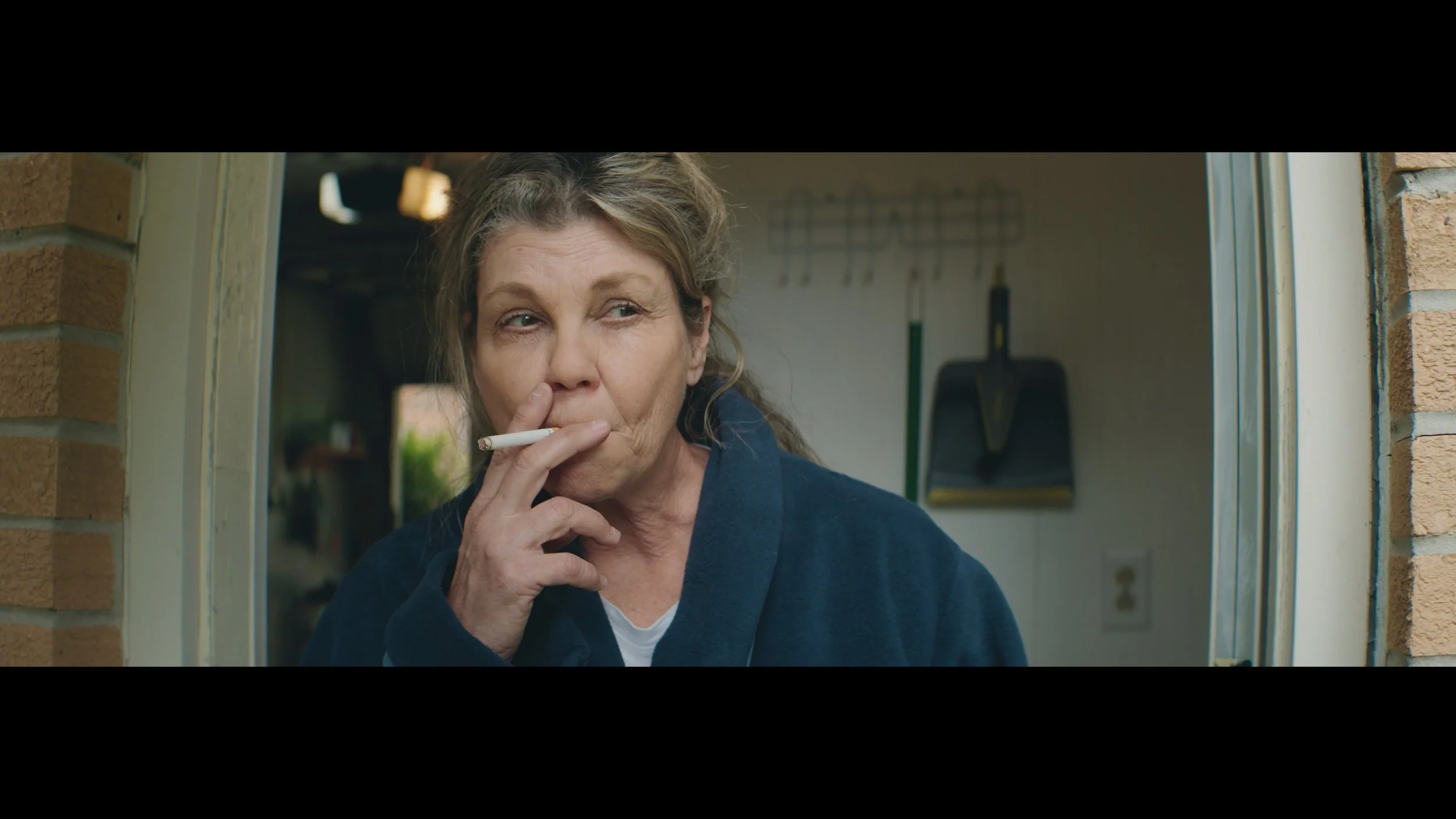 FIRST CONTACT screen capture: Actress Julie Haught is dressed as a weathered woman with serious expression, holding a cigarette near her mouth, standing at a doorway.