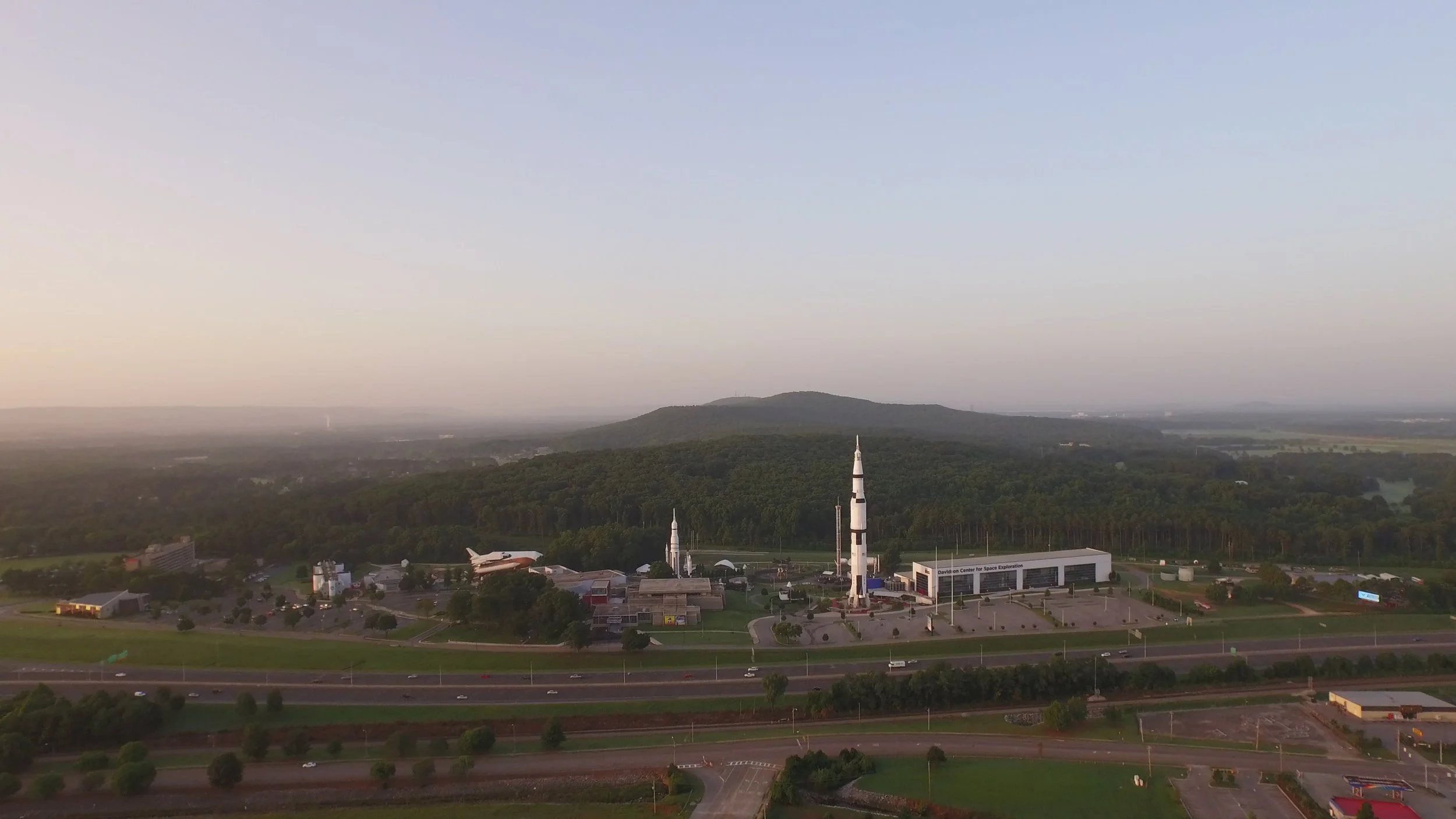 ROCKET CITY SHAKEDOWN screen capture: US Space & Rocket Center, featuring a tall rocket to the right, several launch pads with rockets. There are trees and a parking lot surrounding the facility, with mountains and a wide, open sky in the background.