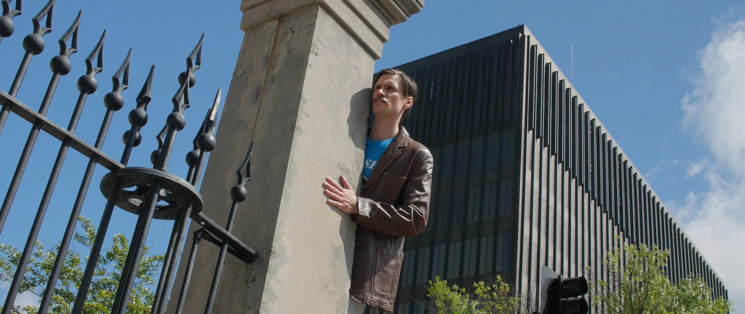ROCKET CITY SHAKEDOWN screen capture: Actor Lincoln Greer stands behind a wrought iron fence, leaning against a stone pillar, observing something. Huntsville court house is visible in the background under a clear blue sky.