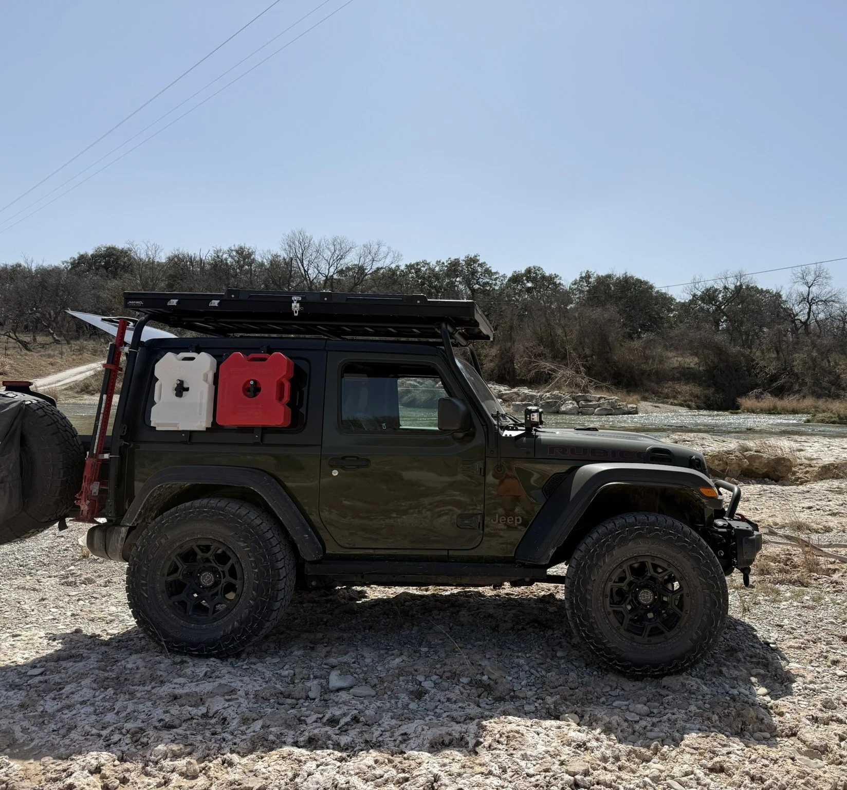 Black Jeep Rubicon parked on rocky terrain near a river with trees in the background, carrying two gas cans on the side and a roof rack.