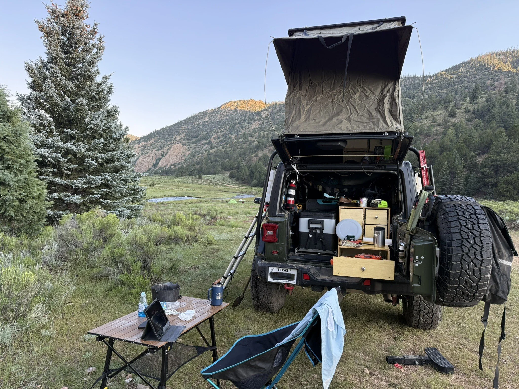Camping setup in a grassy field with a Jeep, open rear hatch containing camping gear, a small table with a laptop, water bottle, and other items, a chair, and a mountain landscape with trees and a river in the background.