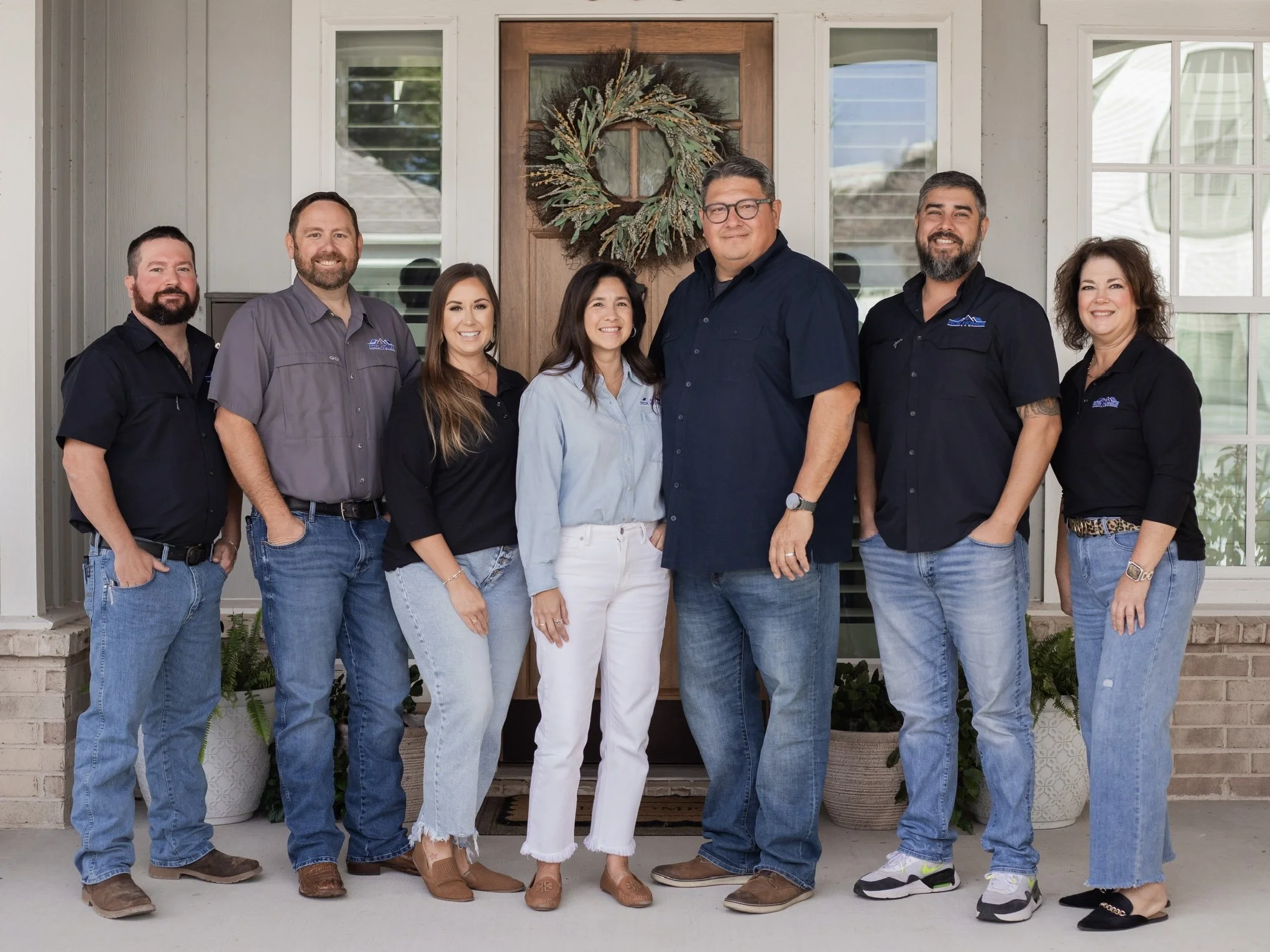 Group of The RCR Homes Team consisting of seven people standing on porch in front of a house with a wooden door decorated with a circular wreath made of greenery and dried flowers.