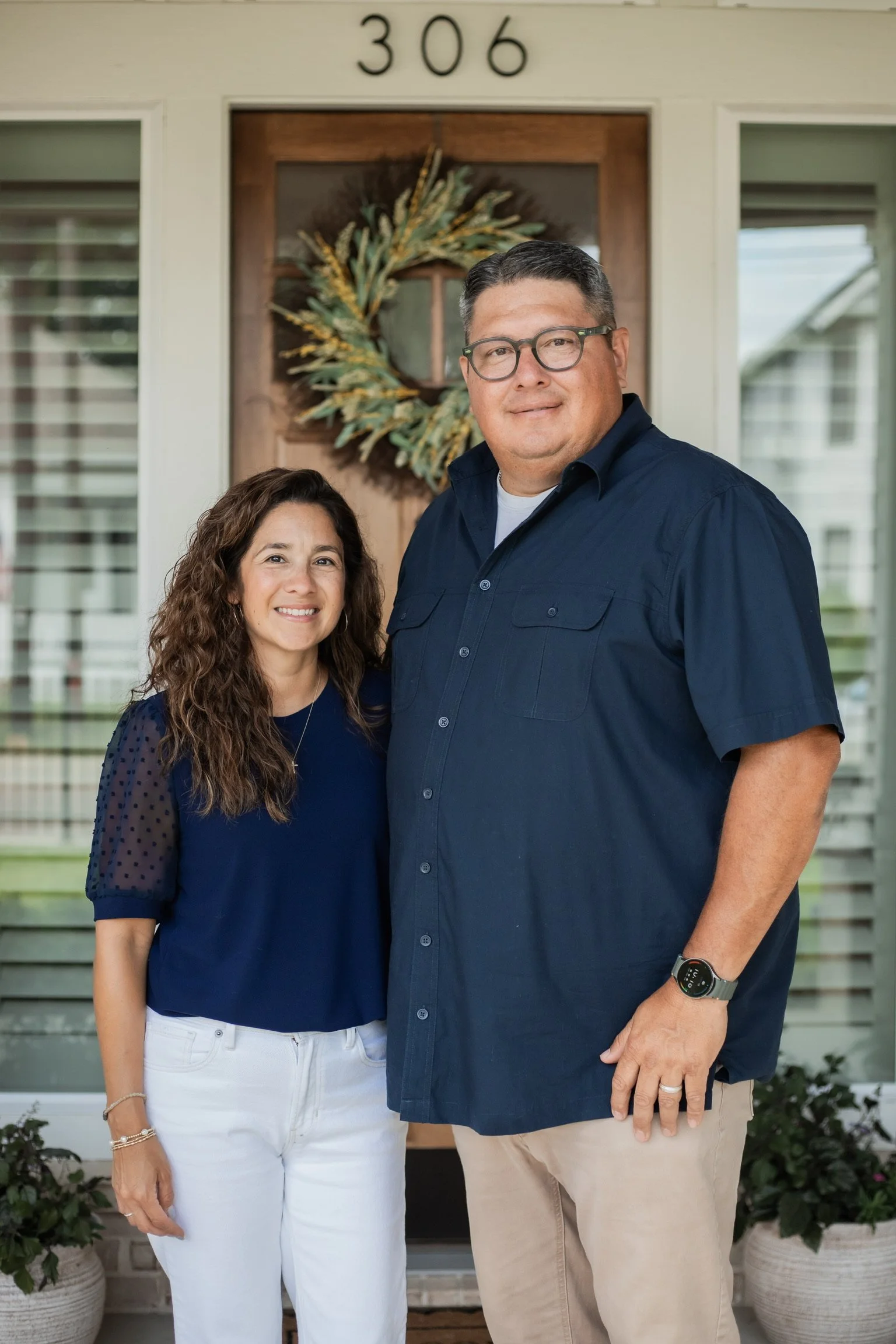 A couple who owns RCR Homebuilders and are standing in front of a house with the number 306 above the door, decorated with a wreath, smiling at the camera.