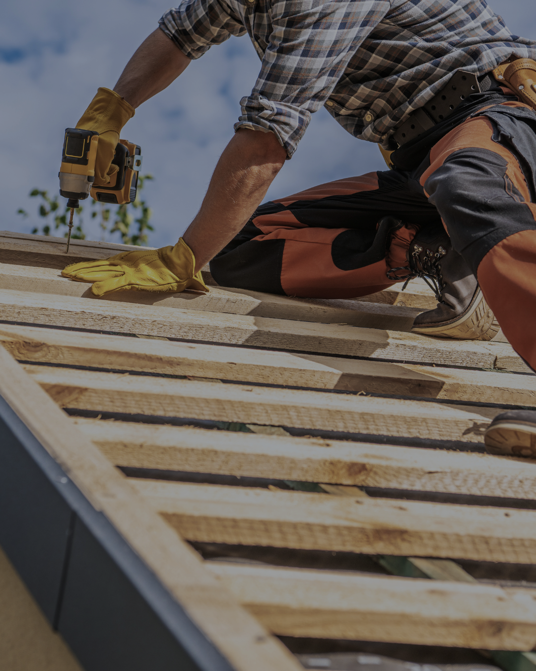 A person in work gloves using a cordless drill working on a roof, wearing an orange and black work pants and a plaid shirt in Victoria, Texas.