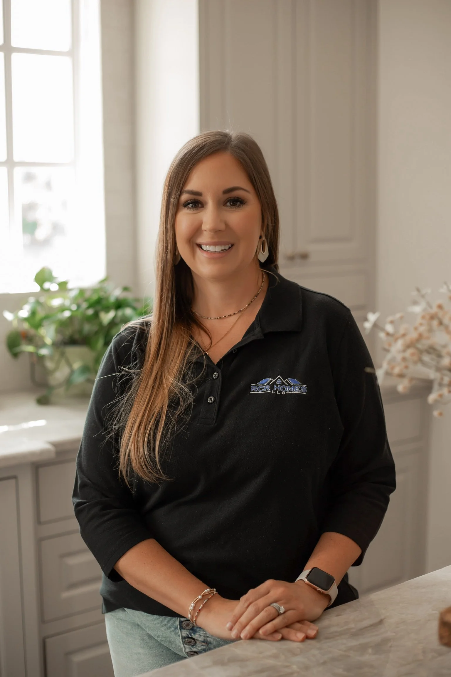 A woman with long blonde hair, wearing a black polo shirt with a logo, standing in a bright kitchen near a window and green potted plants, smiling.