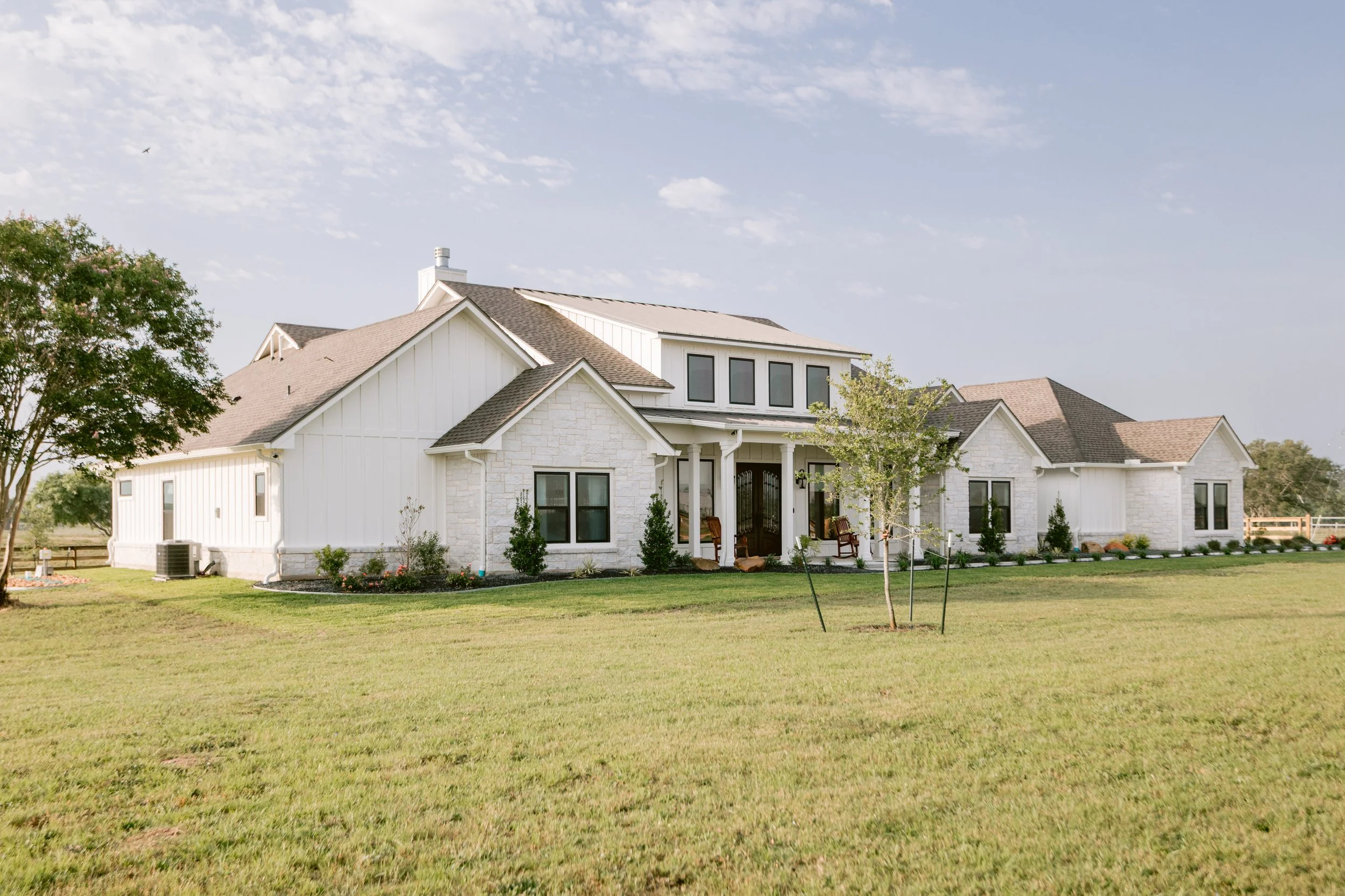 A newly built large white house with multiple gable roofs and a porch, surrounded by a grassy lawn with small trees and shrubs, under a partly cloudy sky in Victoria, Texas.