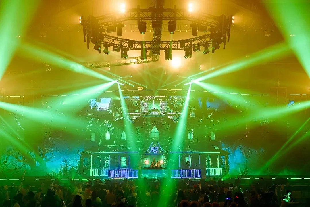 A concert stage with bright green and yellow lights, a large circular lighting rig, and a crowd of people in front.