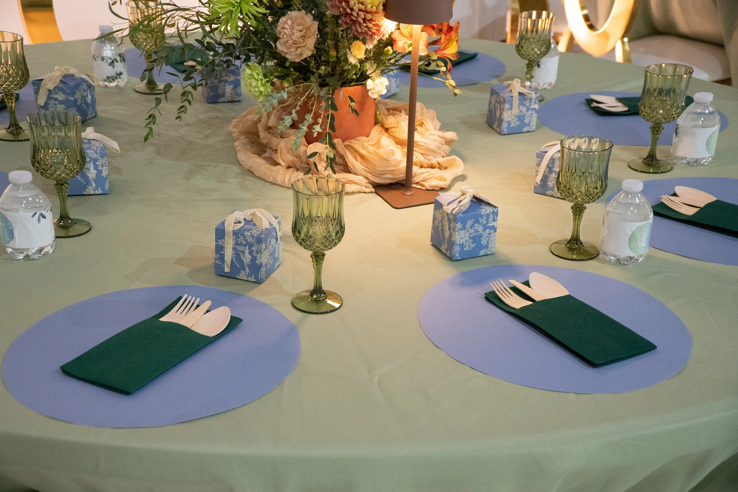 A round dining table set with blue placemats, white napkins, and green napkin holders. There are wine glasses and water bottles placed around the table. A centerpiece with flowers and a lamp is in the middle of the table.