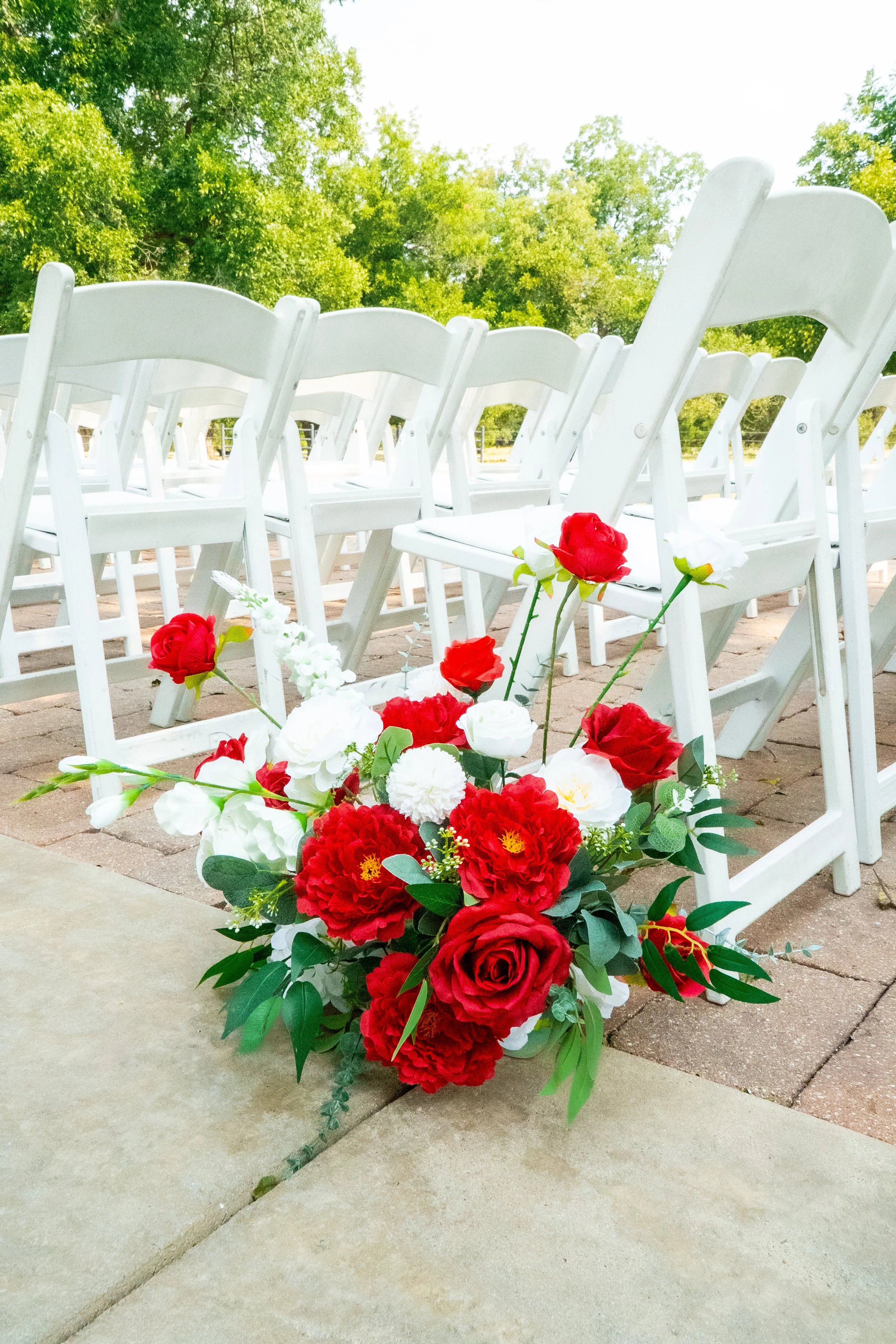 A bouquet of red, white, and green flowers arranged on a concrete surface in front of empty white chairs at an outdoor ceremony setup with green trees in the background.