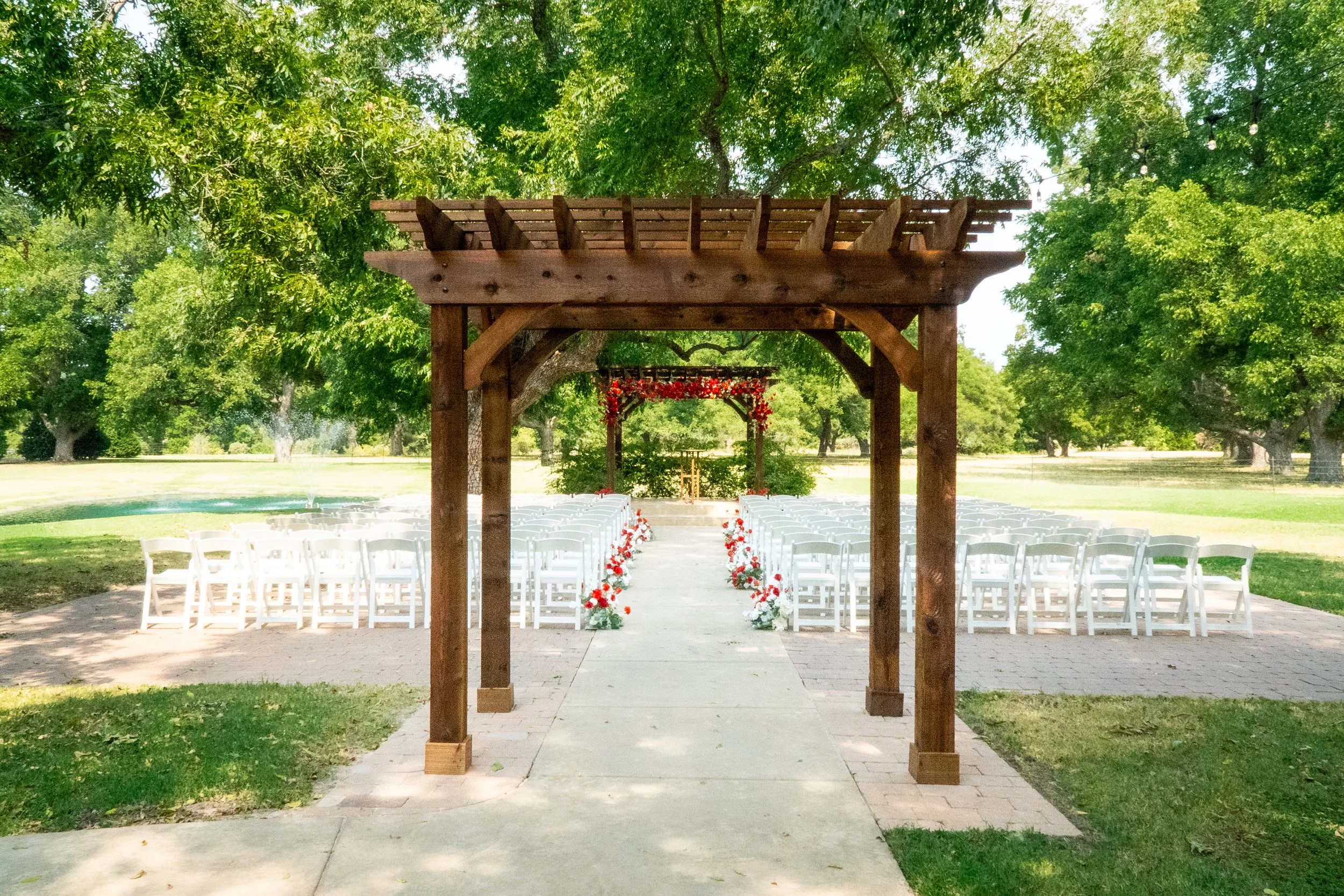 Outdoor wedding setup with white chairs arranged in rows on either side of an aisle with red and white flower decorations, under a wooden arbor, in a park with green trees and a fountain in the background.