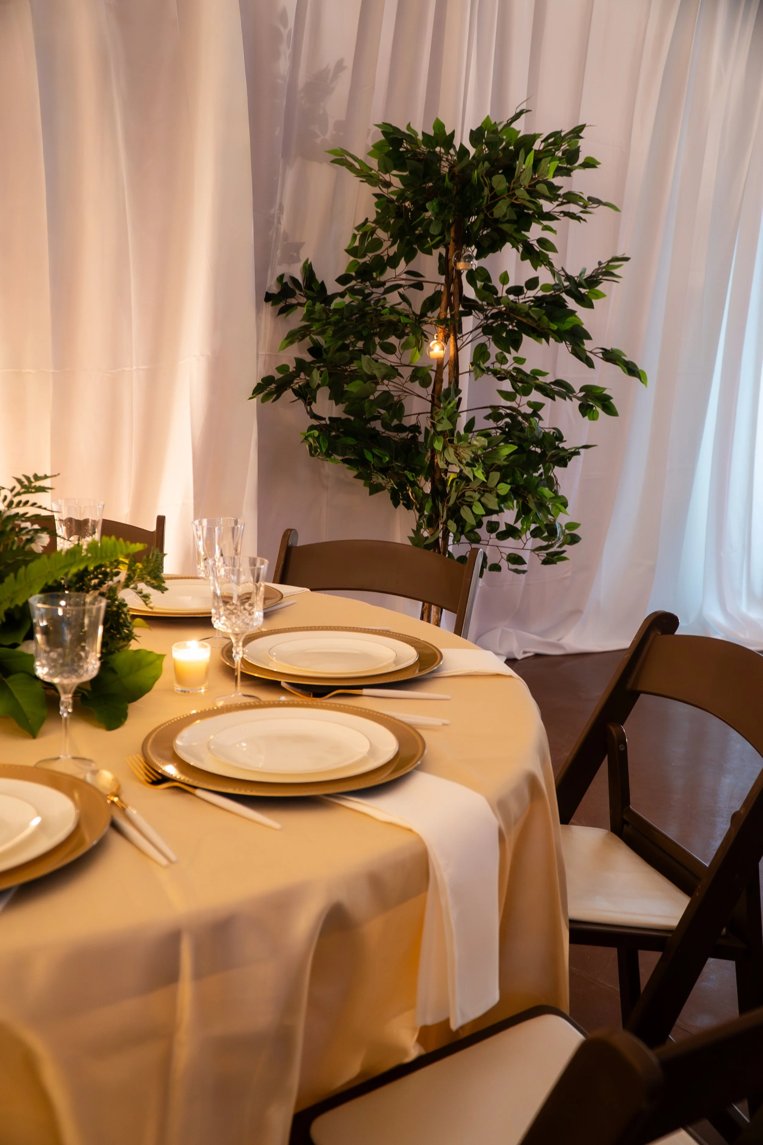 Decorated dining table with white tablecloth, gold-rimmed plates, wine glasses, silverware, and green foliage centerpiece. A tall, leafy green plant with string lights is in the background, next to white curtains.