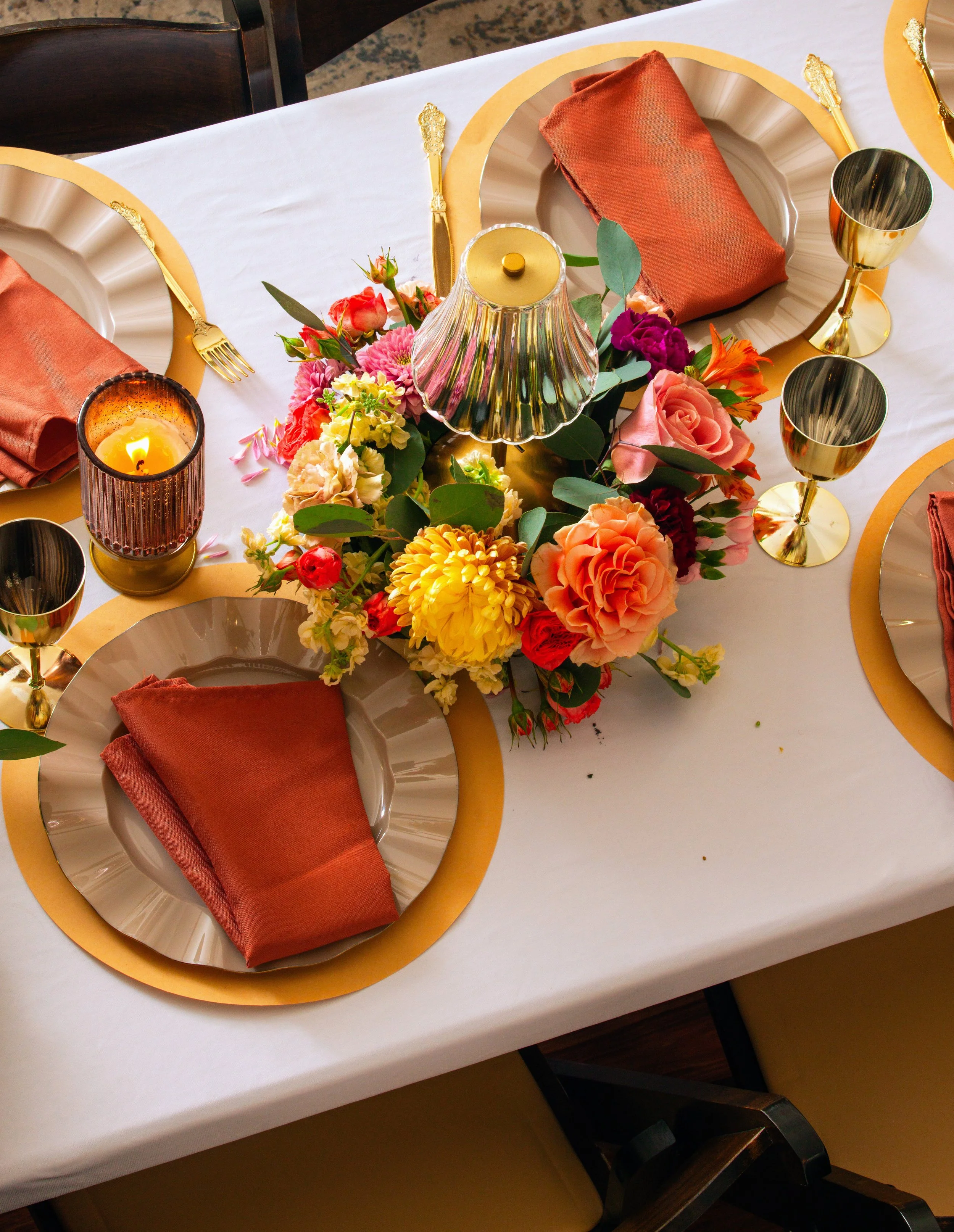 A table with a white tablecloth, set for a formal dinner with gold-rimmed plates, copper cloth napkins, gold utensils, gold-rimmed glasses, a floral centerpiece with various colorful flowers, a small lamp, and a lit candle.