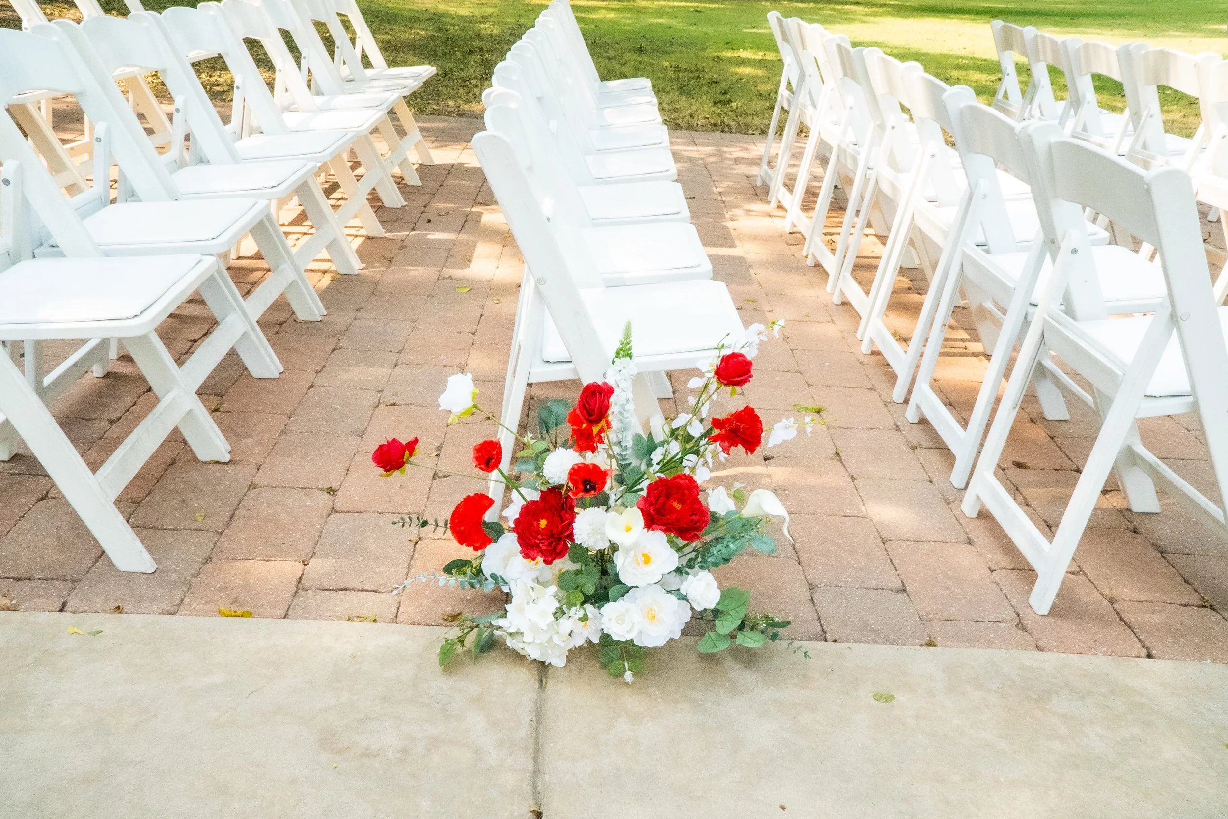 Outdoor wedding or special event setup with white folding chairs arranged on brick pavers, facing a floral arrangement of red, white, and green flowers, set against a grassy background.