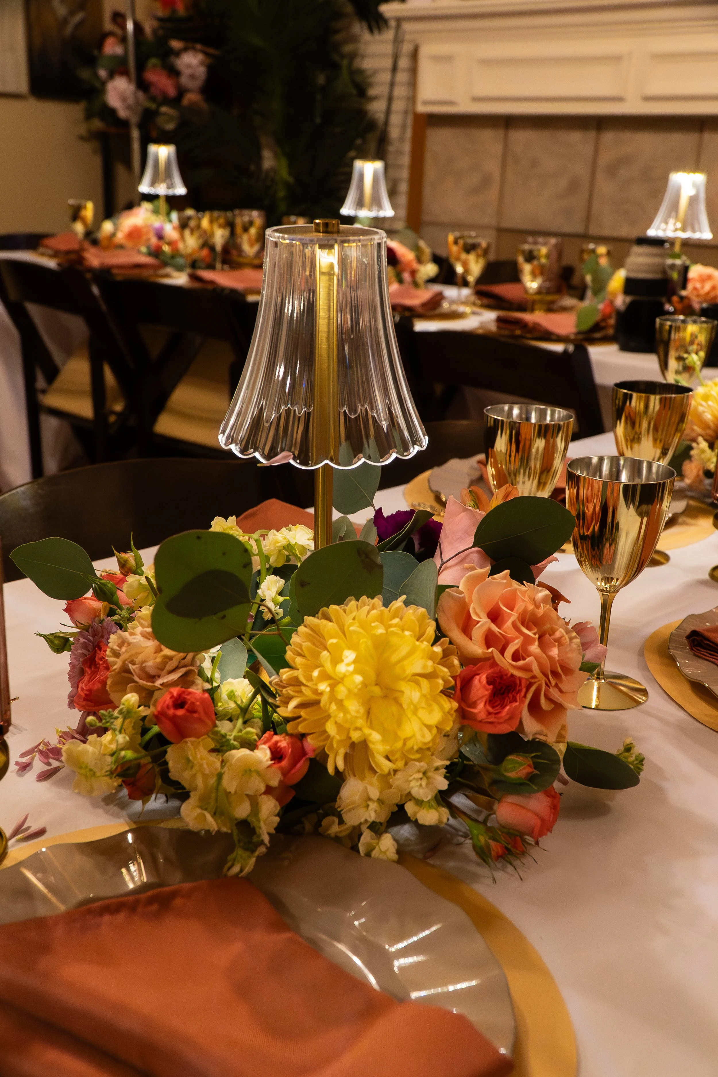 Elegant table setting with a floral centerpiece, gold and glass goblets, a small lamp, and neatly folded cloth napkins, for a formal event or celebration.
