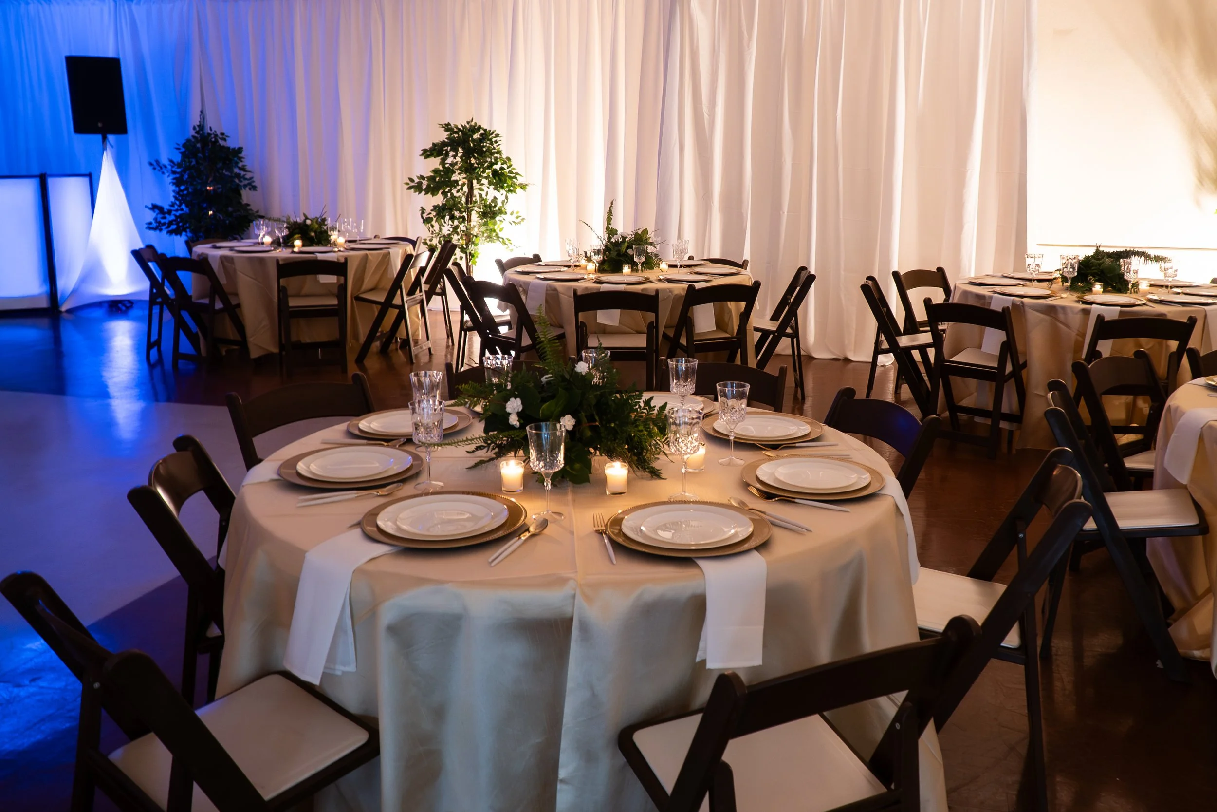 Round banquet tables with white tablecloths, elegant dinnerware, candles, and floral arrangements, set for a formal event in a decorated event space with draped white curtains and ambient lighting.