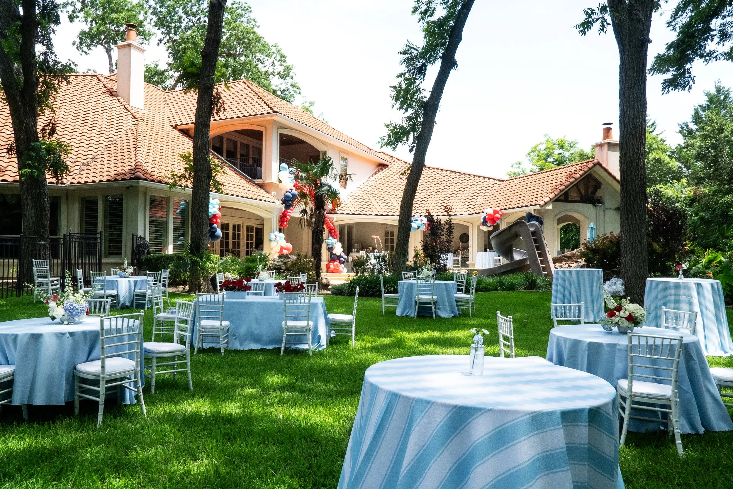 A backyard with round tables covered in light blue tablecloths, decorated with floral centerpieces, set up for an outdoor event in front of a large house with a red tile roof and surrounded by trees.