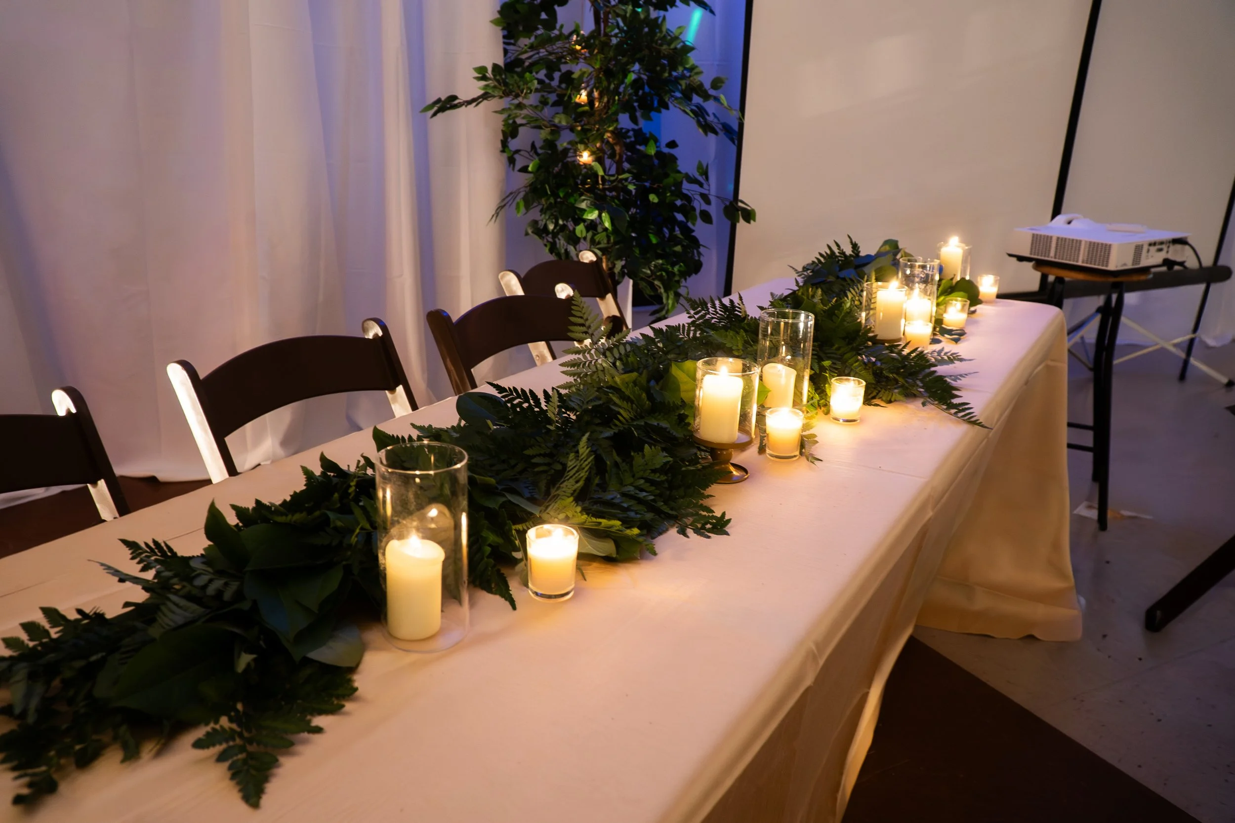A long banquet table decorated with candles and green foliage, set in a dimly lit room with white drapery and a large leafy plant in the background.
