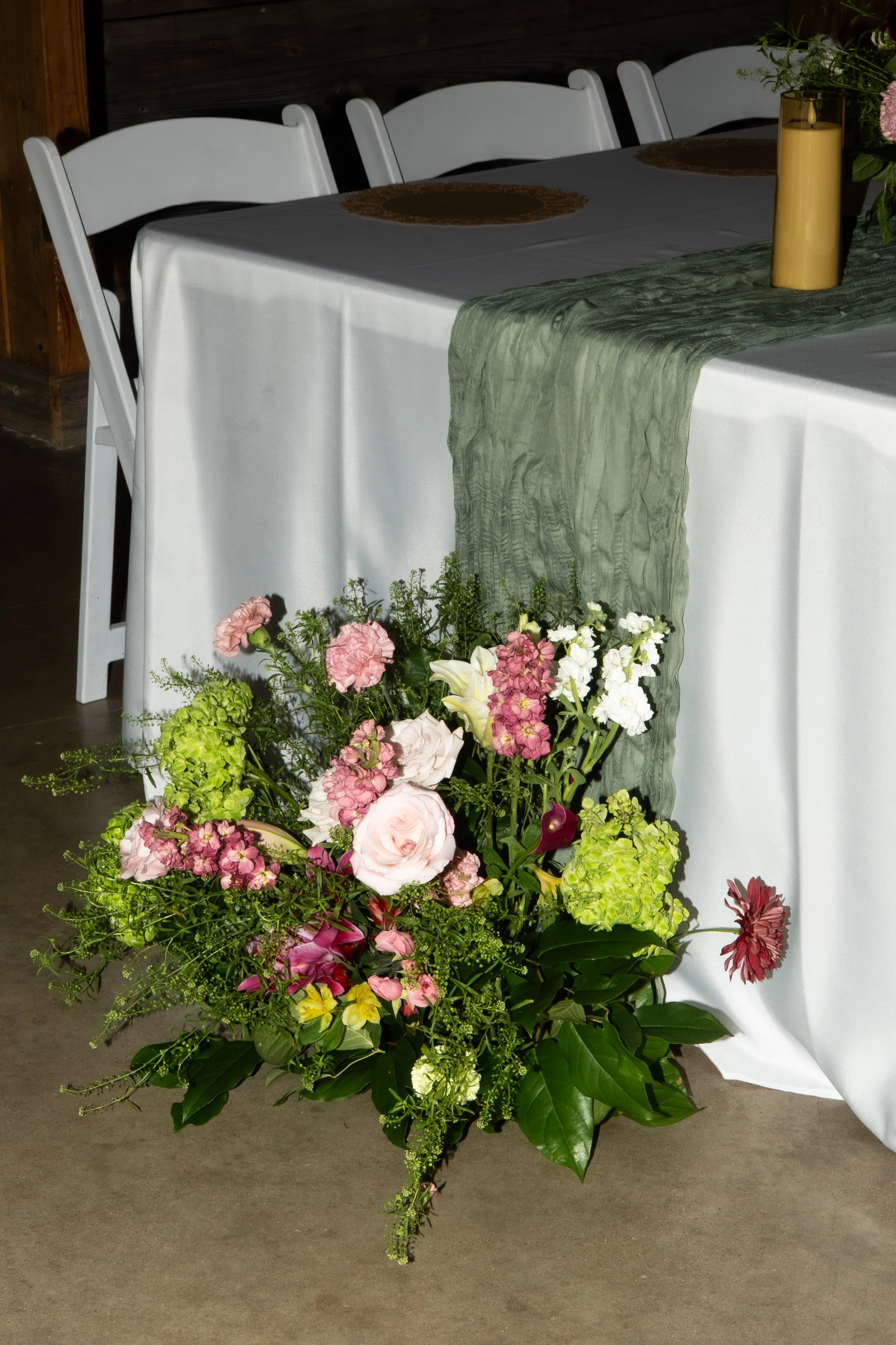 A floral arrangement of pink, white, and green flowers placed at the corner of a decorated table with a white tablecloth, green runner, and vase with pink flowers inside.