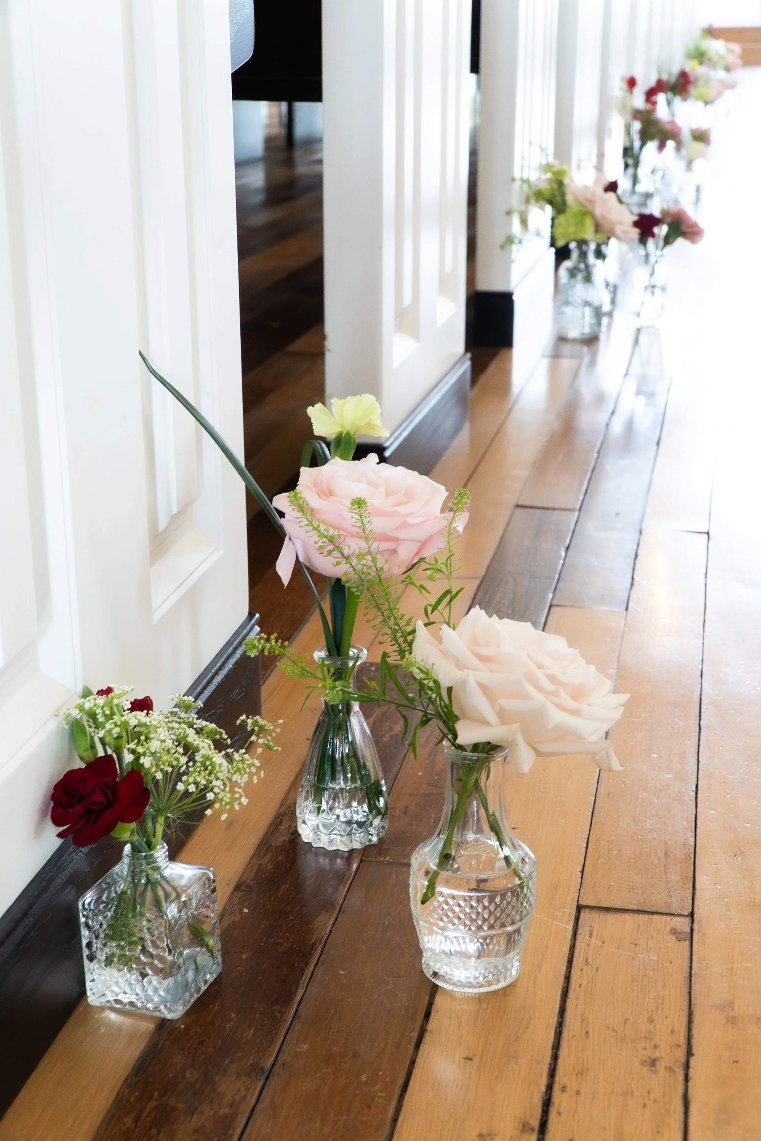 Three small glass vases with flowers on a wooden floor near a white panel door. The vases contain pink, white, and red flowers.