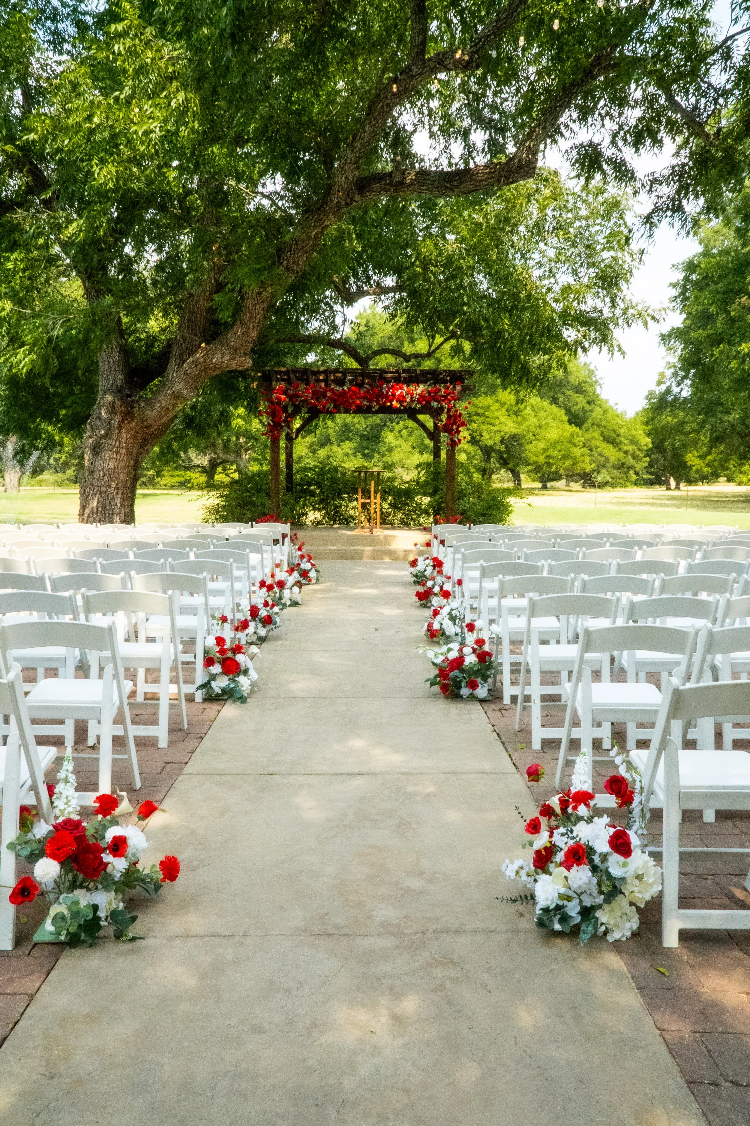 Outdoor wedding ceremony setup with white chairs arranged on either side of a central aisle, decorated with red, white, and green floral arrangements. An arbor decorated with red flowers stands at the altar under a large tree with lush green leaves, 