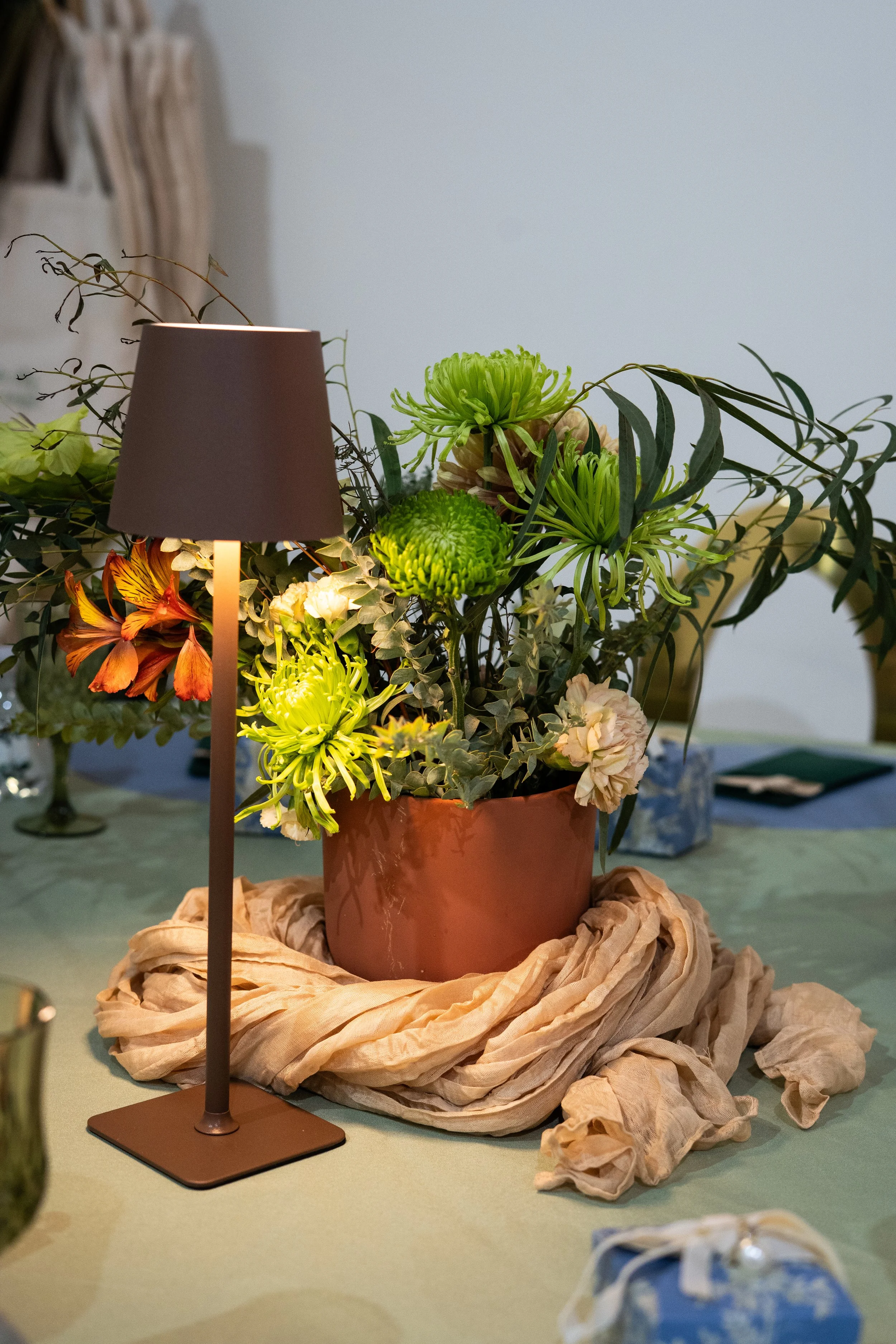 A floral arrangement in a reddish-brown pot on a table, surrounded by beige fabric and other tableware, with a brown lamp partially blocking the view of the flowers and a small wrapped gift on the table.