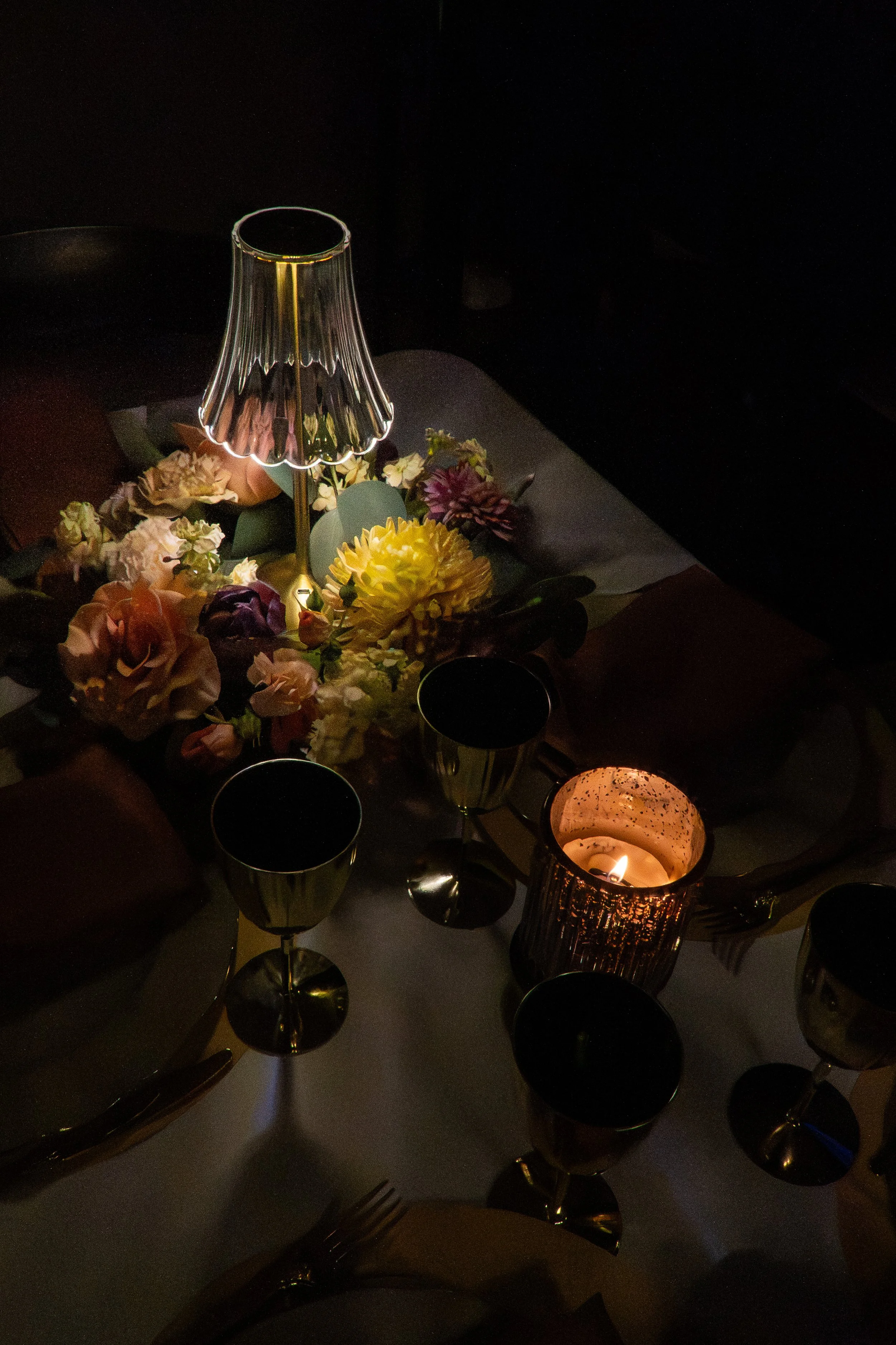 A dimly lit table setting with a small lampshade, candles, wine glasses, and a floral centerpiece. Fort worth wedding
