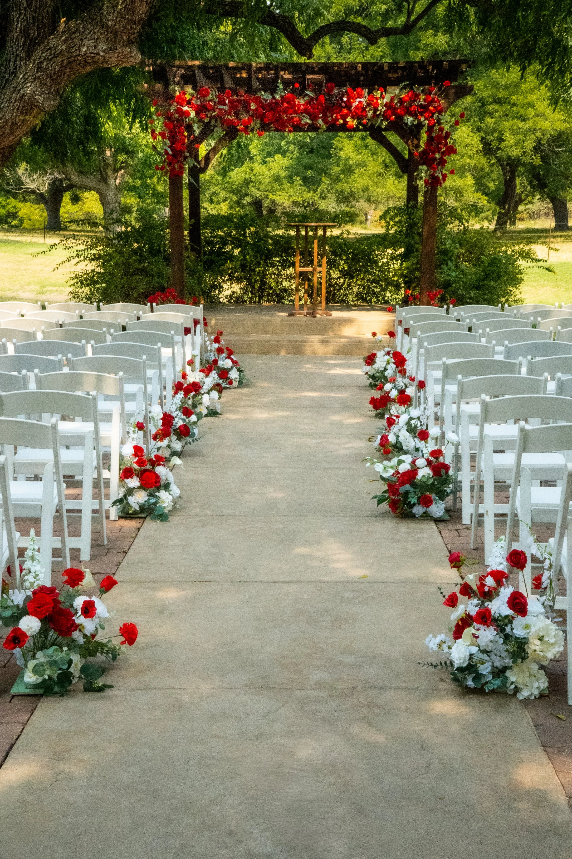 Outdoor wedding ceremony setup with white chairs on either side of a concrete aisle, decorated with red and white flowers, leading to a wooden arch draped with red flowers, set in a park with trees and greenery.