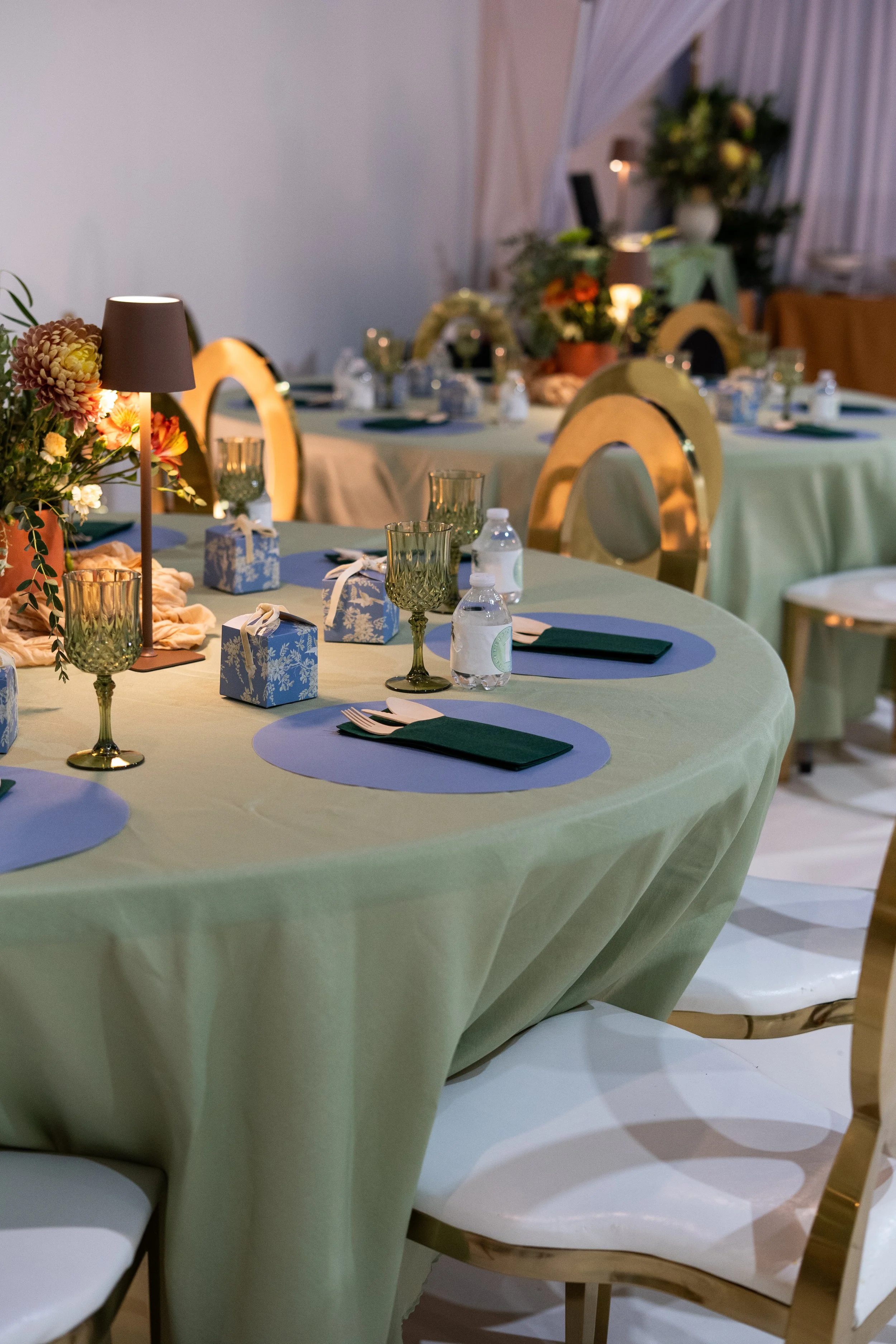 A decorated dining table with a light green tablecloth, green and gold glasses, small gift boxes, and floral centerpieces with candles in a softly lit room with white curtains.