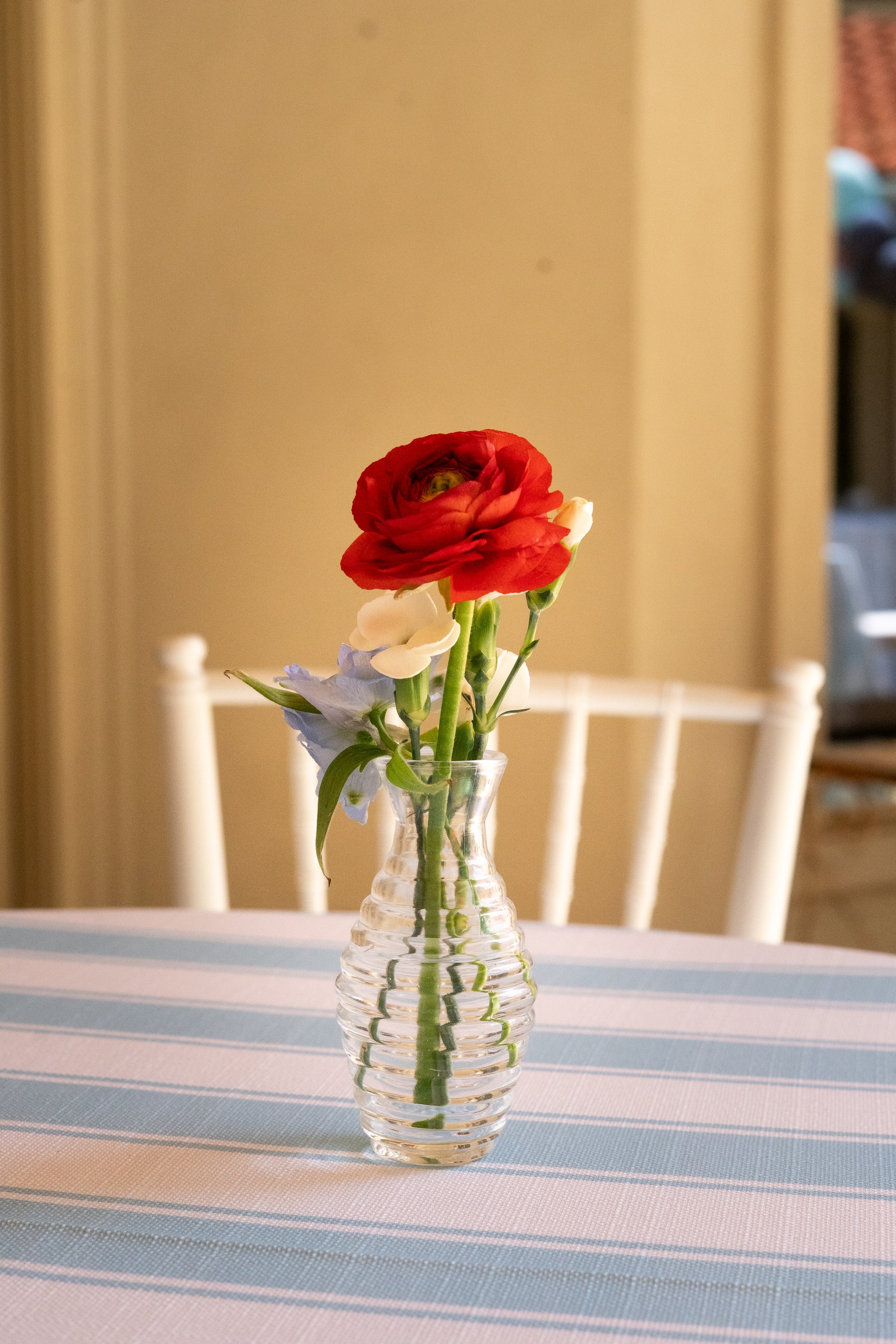 Red and white flowers in a clear ribbed glass vase on a table with a blue and white striped tablecloth, in a room with a beige wall and beige chairs.