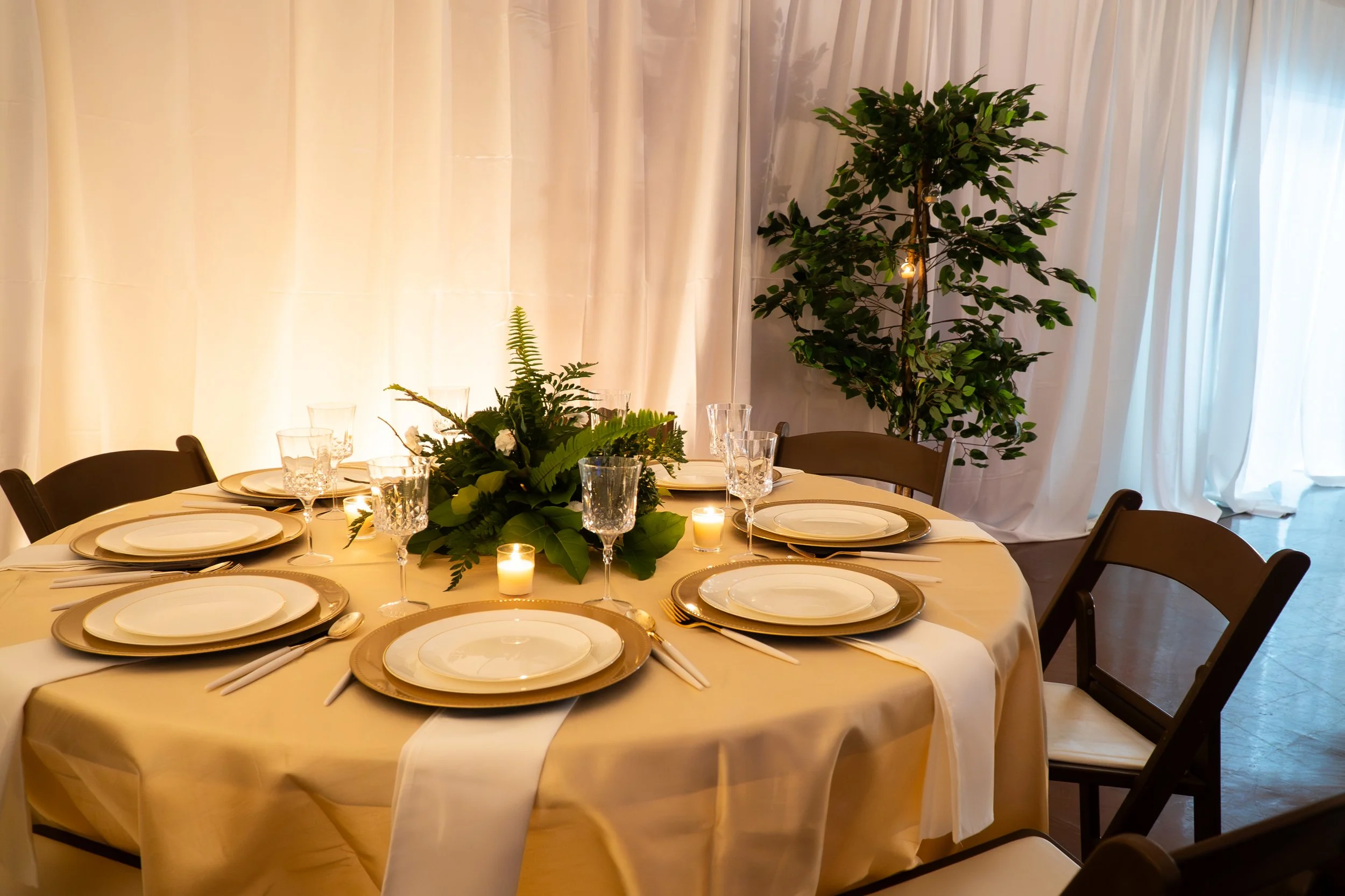 Round banquet table set with white napkins, white plates with gold rims, crystal glasses, and small candles, decorated with a green floral centerpiece. The background features cream-colored curtains and a large potted plant.