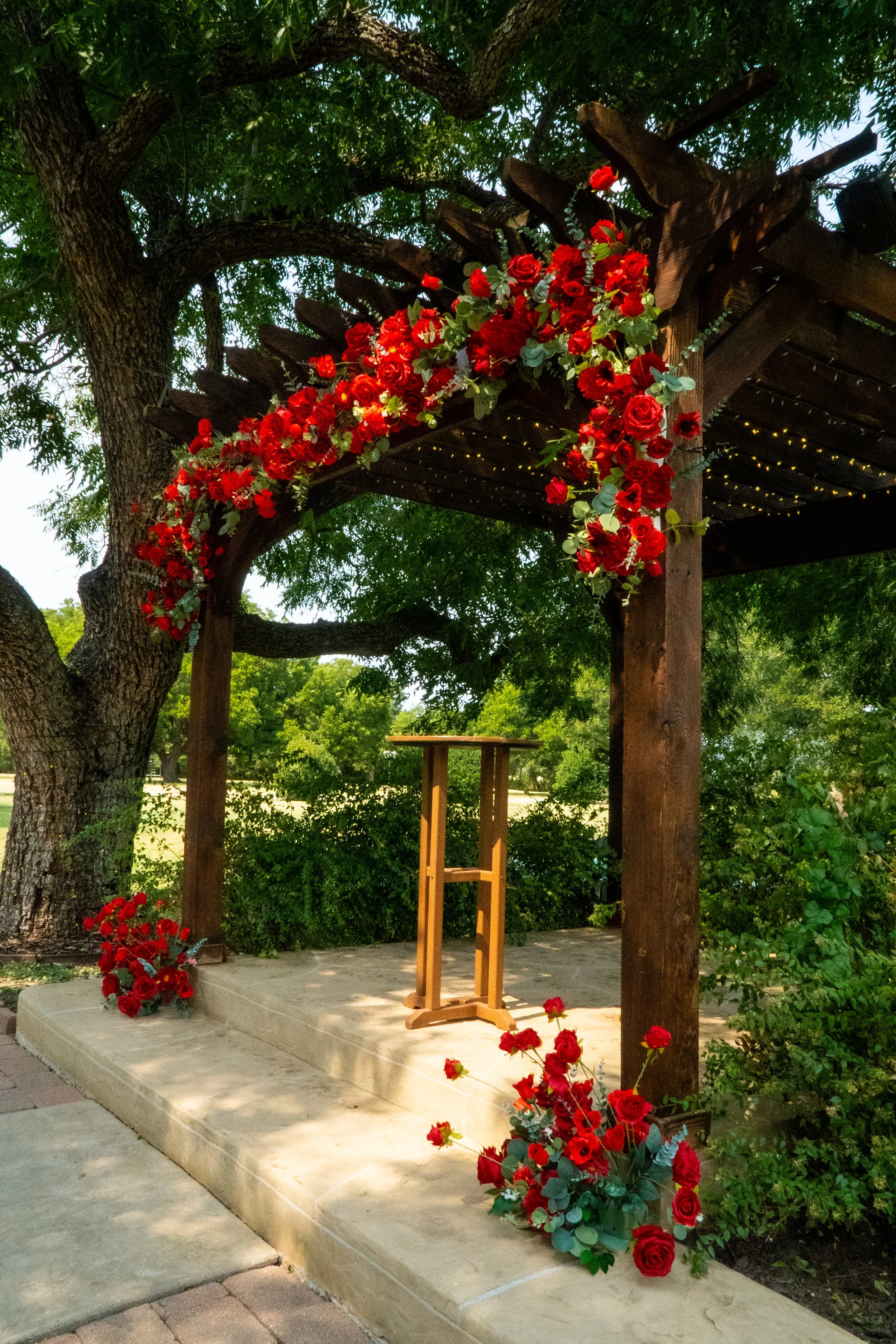 Wooden wedding arch decorated with red roses and greenery, set outdoors under a large tree with green leaves.